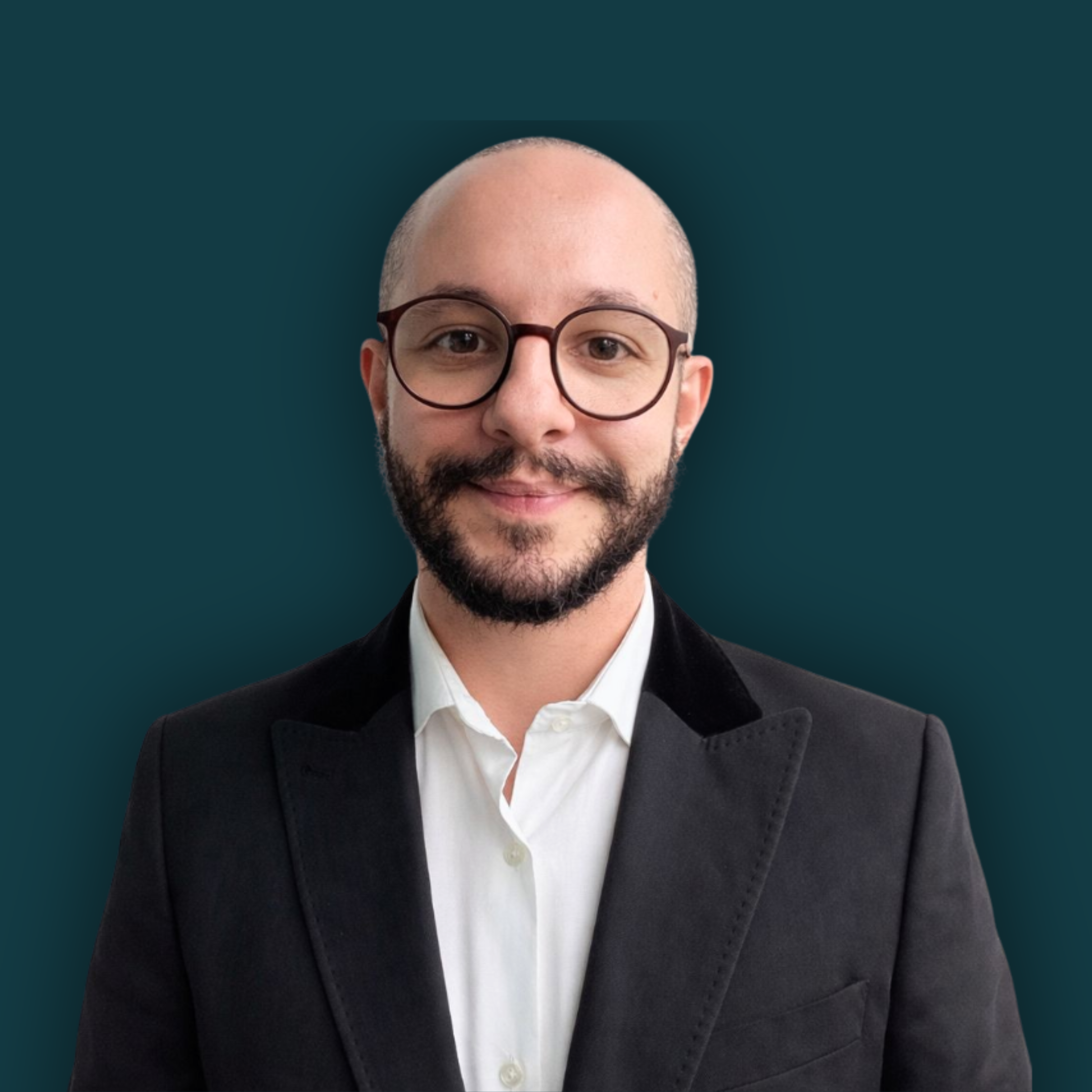 A portrait of a man with glasses and a beard, dressed in a white shirt and black blazer, smiling against a dark teal background.