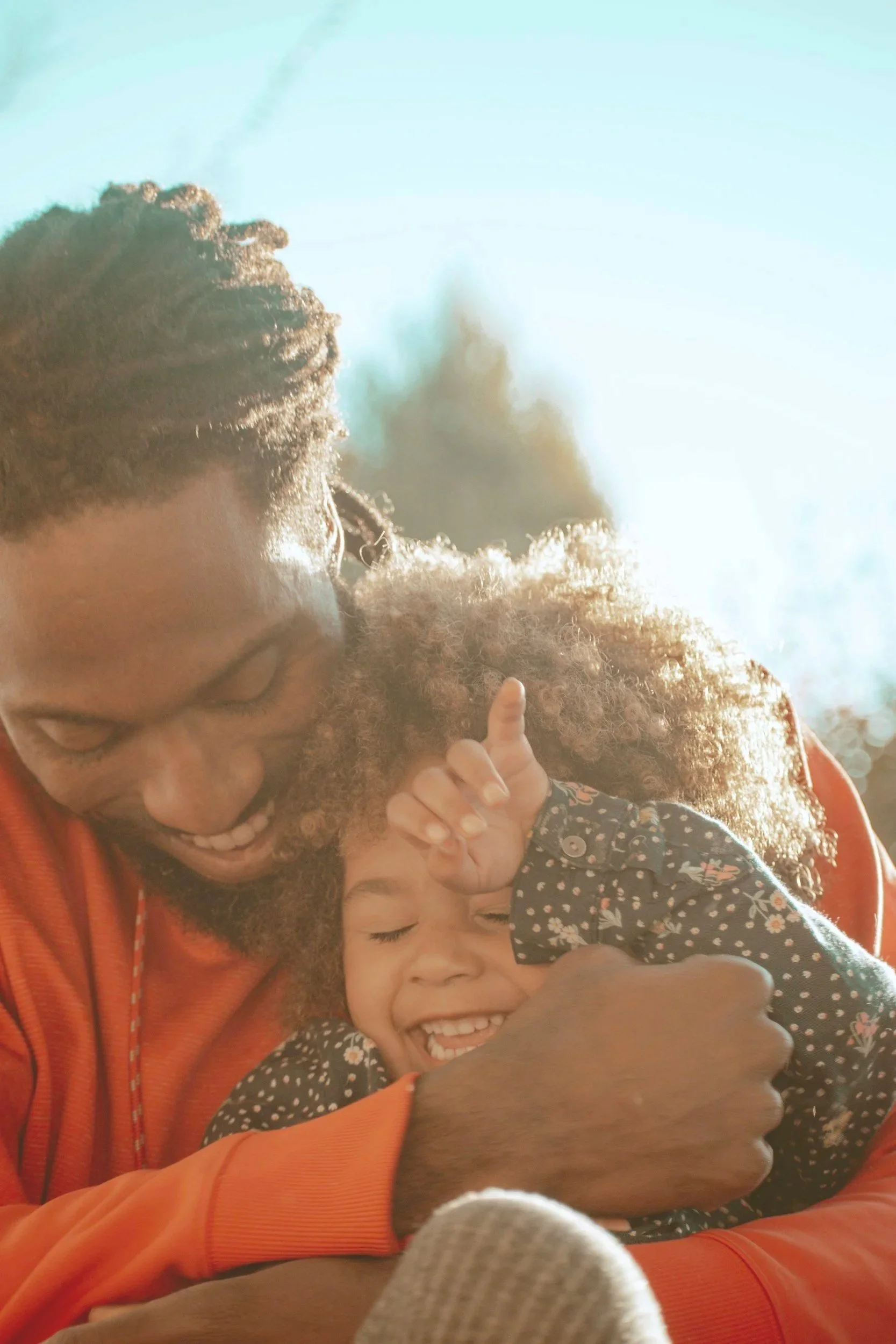 A man with glasses and curly hair smiling and hugging two children, one of whom is a girl with dark hair and a floral dress, outdoors with sunlight in the background.