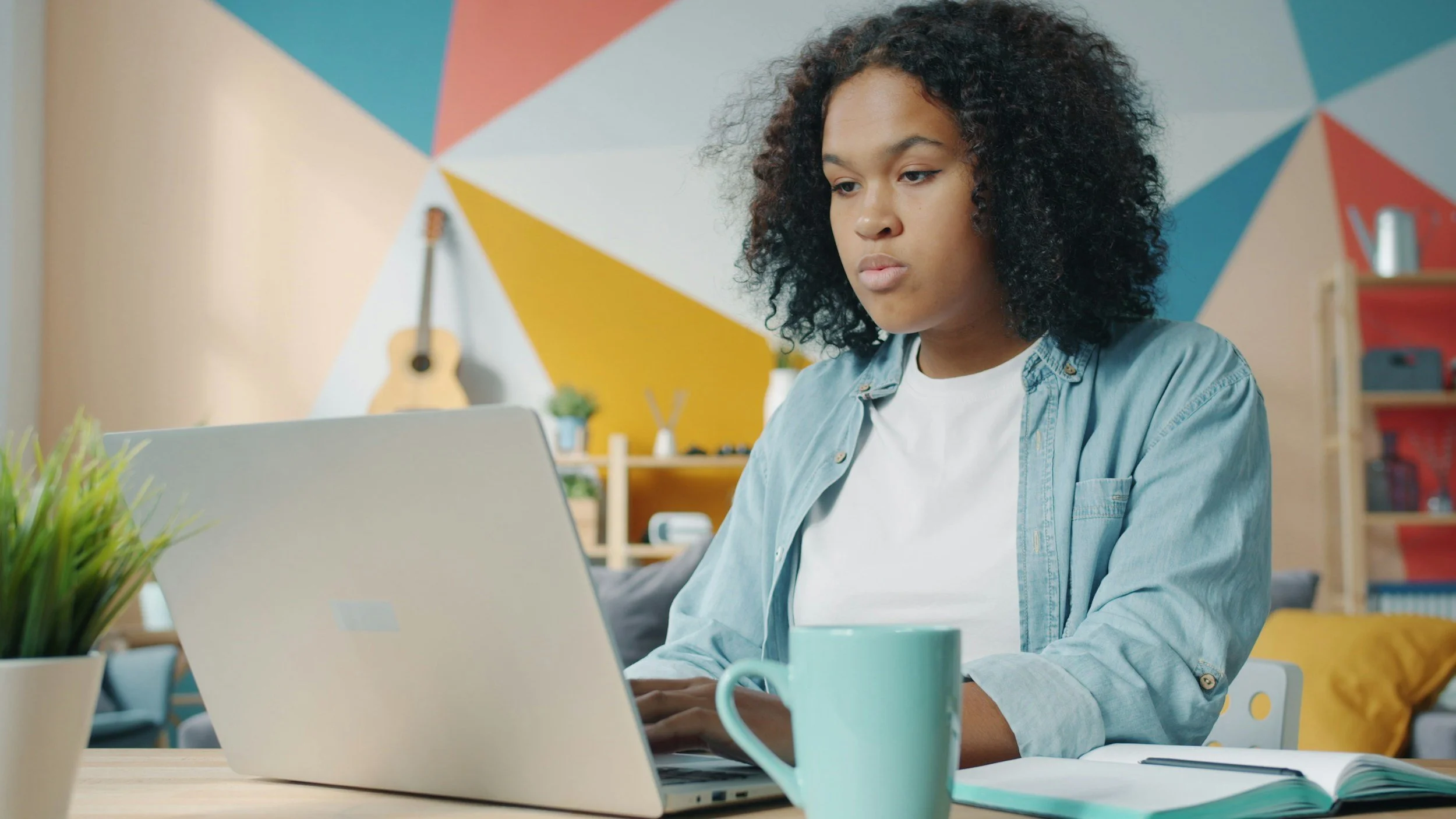 A woman with curly hair working on a laptop at a desk surrounded by a mug, plant, and open notebook, in a colorful modern room.