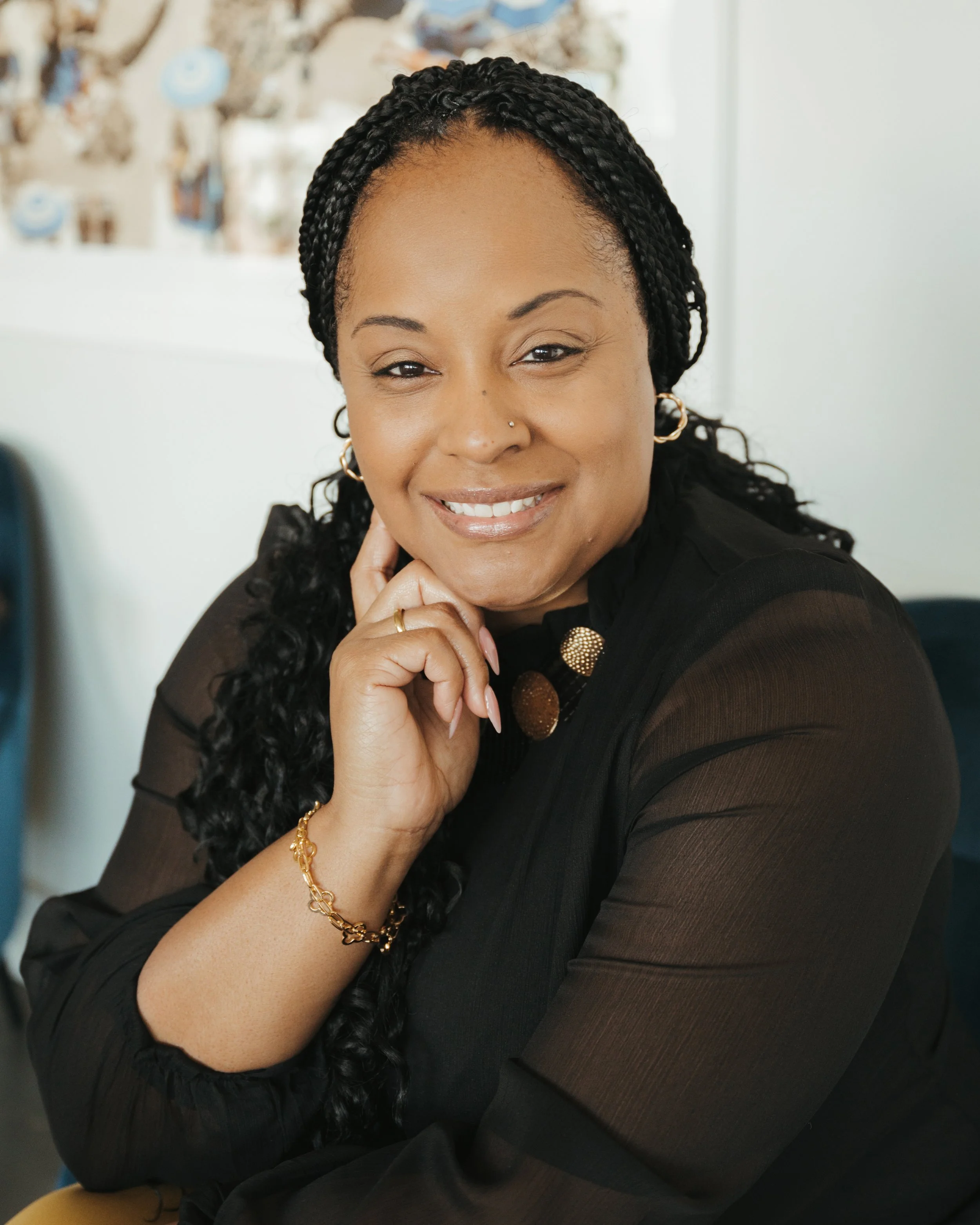 A smiling woman with long braided hair, wearing a black sheer top, gold jewelry, and earrings, resting her chin on her hand in an indoor setting.
