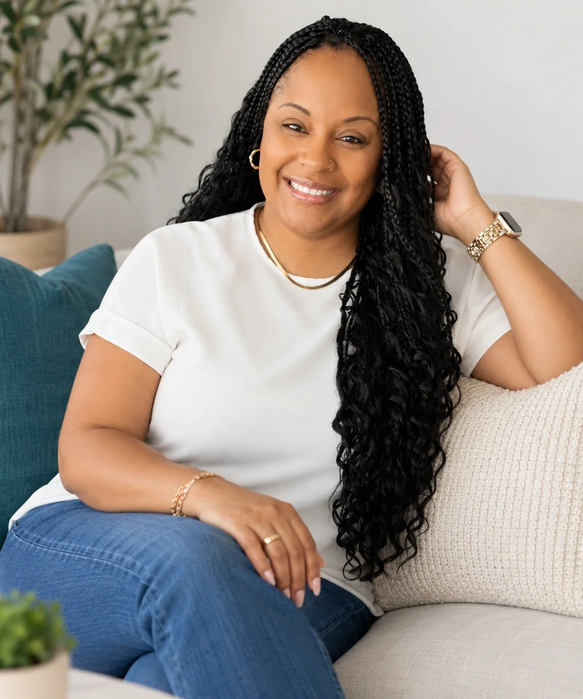 Smiling woman with long, curly black hair sitting on a beige sofa in a living room, wearing a white t-shirt, blue jeans, gold jewelry, and a smartwatch.