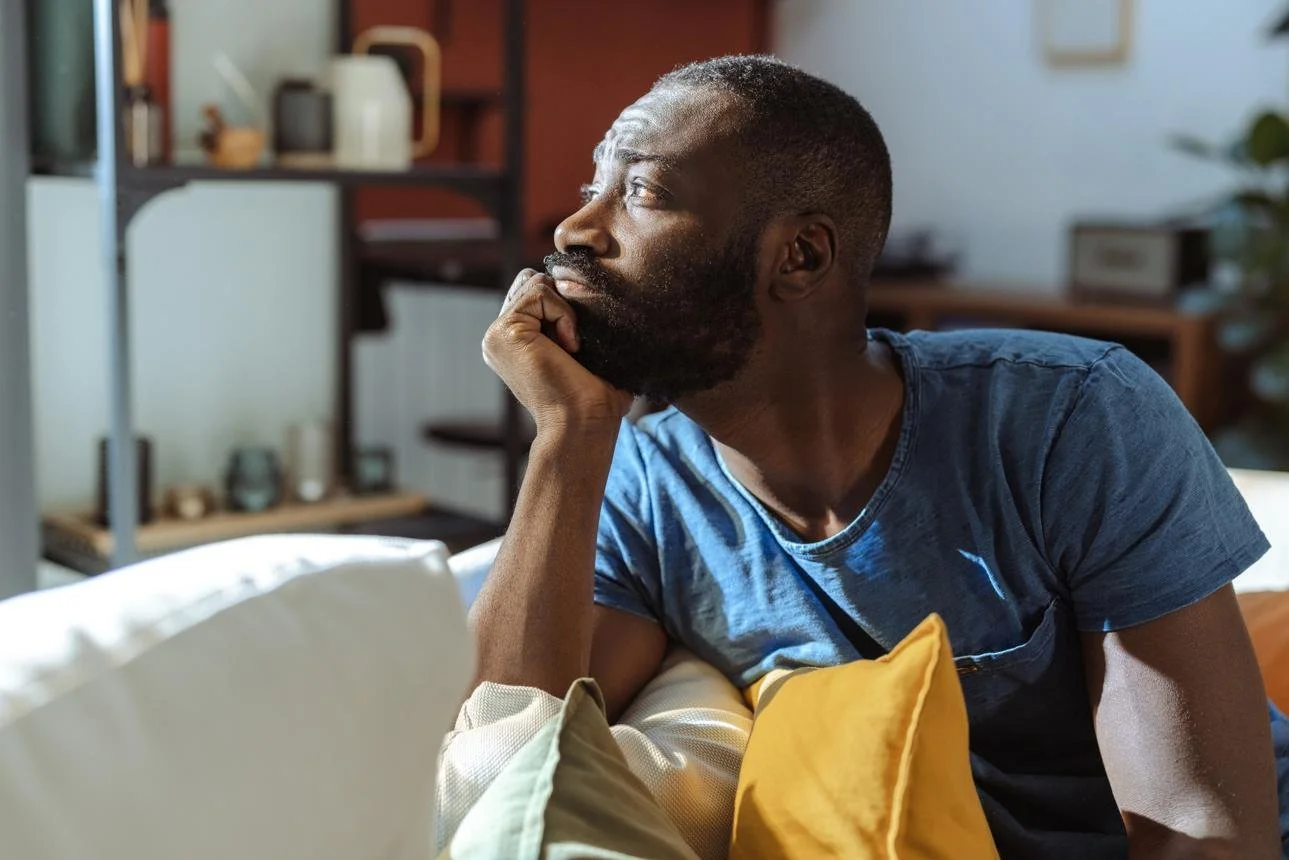 A man with dark skin and a beard sitting on a couch, resting his chin on his hand, looking to the side with a thoughtful expression, in a living room with shelves and decorative items in the background.