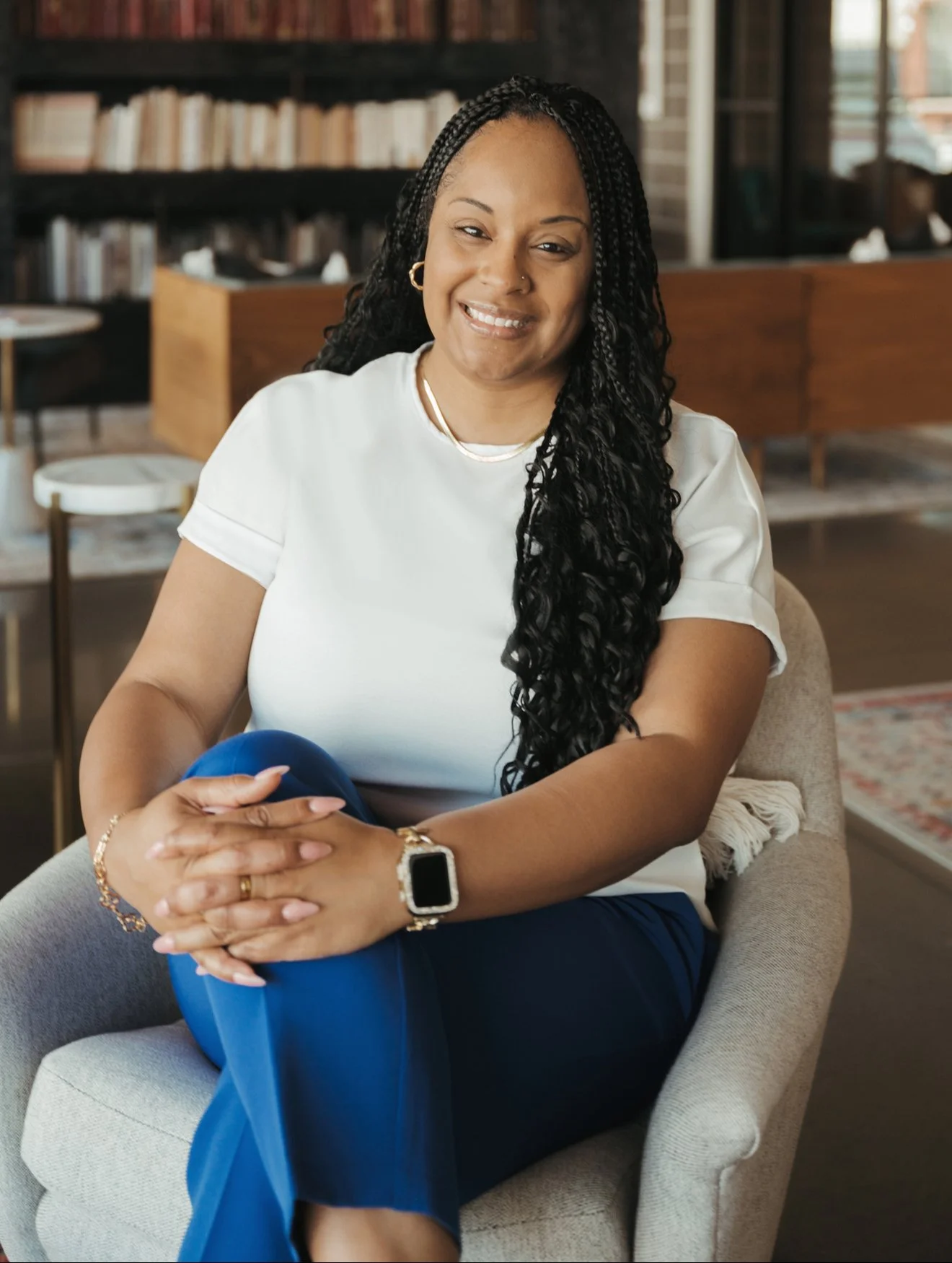 A woman with long black braids, wearing a white t-shirt and blue pants, sitting on a beige armchair in a cozy library setting with bookshelves behind her. She is smiling at the camera with her hands clasped on her knee, wearing a smartwatch, gold hoop earrings, and a gold necklace.