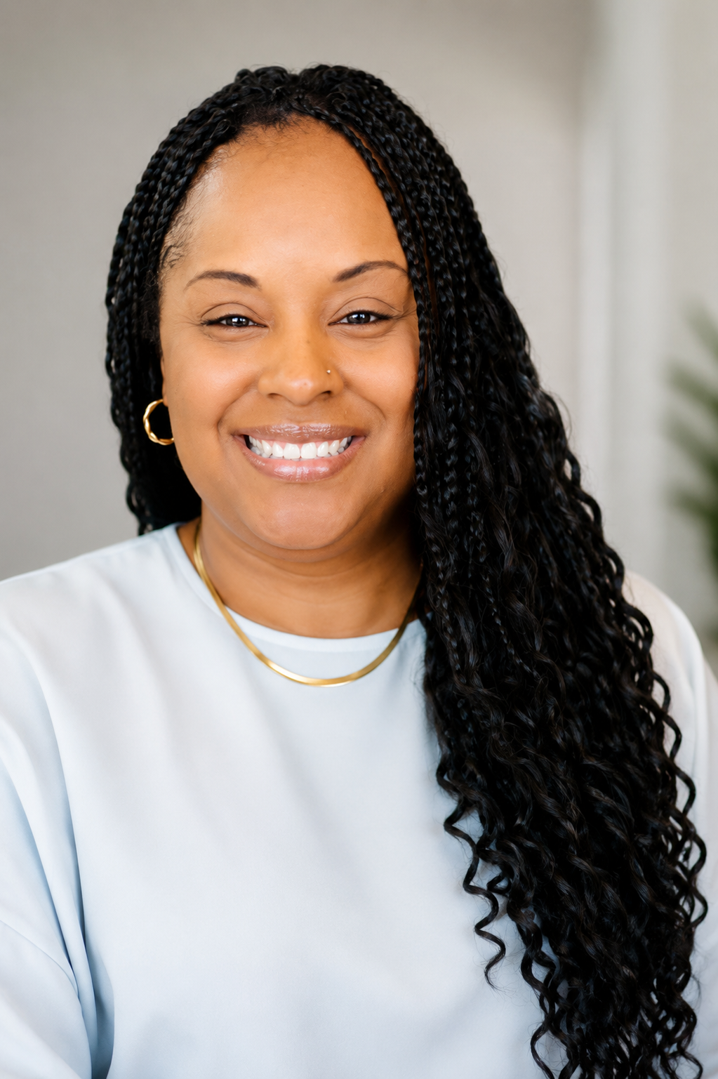 A woman with long braided black hair, wearing a light blue top, gold hoop earrings, and a gold necklace, smiling at the camera.