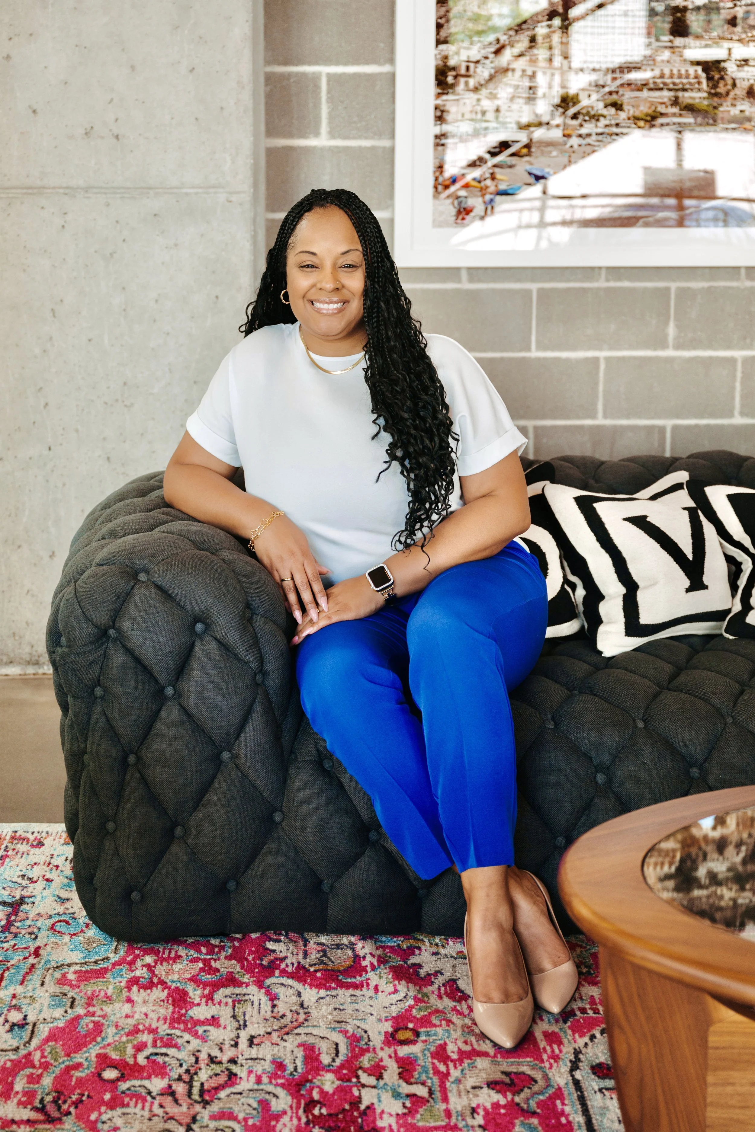 A woman with long, curly black hair is sitting on a dark gray, tufted sofa with a quilted pattern in a modern living room. She is smiling, wearing a white short-sleeve top, royal blue pants, and beige high heels. She has a gold bracelet, a thin gold necklace, and a watch on her wrist. Behind her, there is a large colorful painting on an exposed concrete and brick wall. To her side, there’s a lace-patterned area rug and a wooden side table.