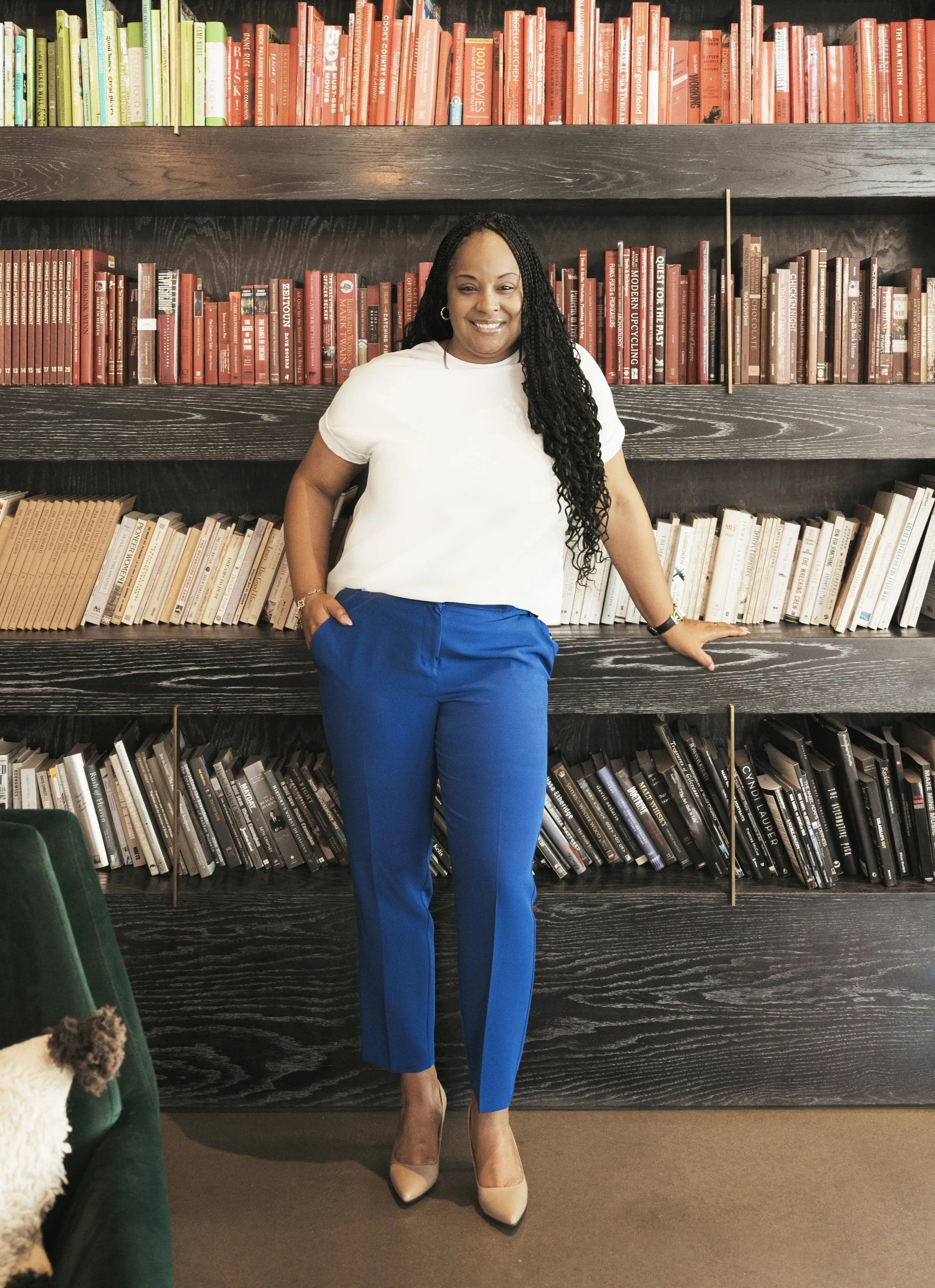 A woman with long curly black hair standing in front of a bookshelf filled with colorful books. She is wearing a white t-shirt, bright blue pants, and beige high heels, smiling at the camera with one hand in her pocket and the other resting on the shelf.