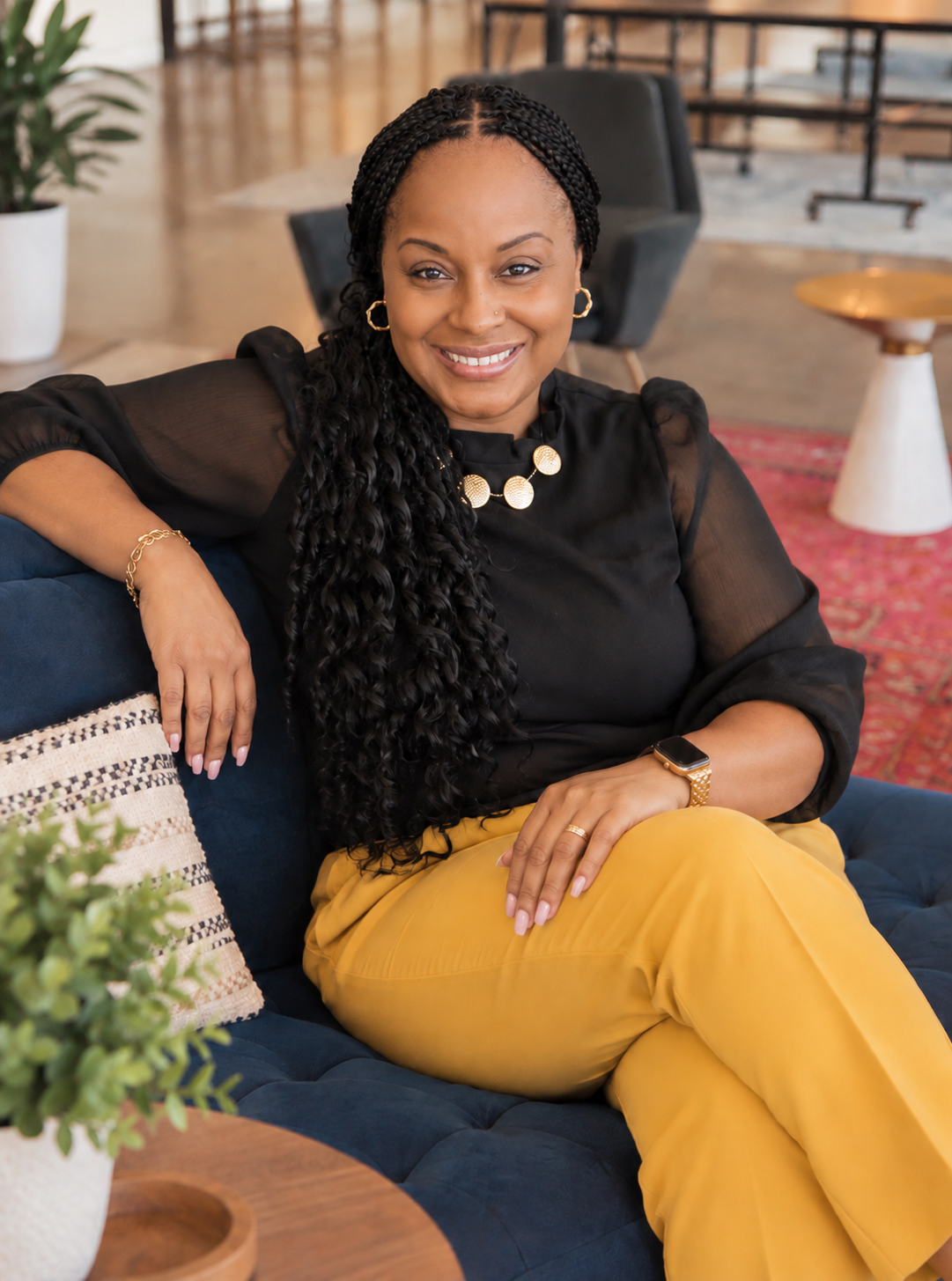 A woman sitting on a dark blue couch, smiling, with her arm resting on the back of the couch. She has long, curly black hair, is wearing a black sheer blouse, yellow pants, gold jewelry including earrings, necklace, bracelet, and watch. The background shows a modern, stylish room with a plant, a red rug, and a small white and gold side table.