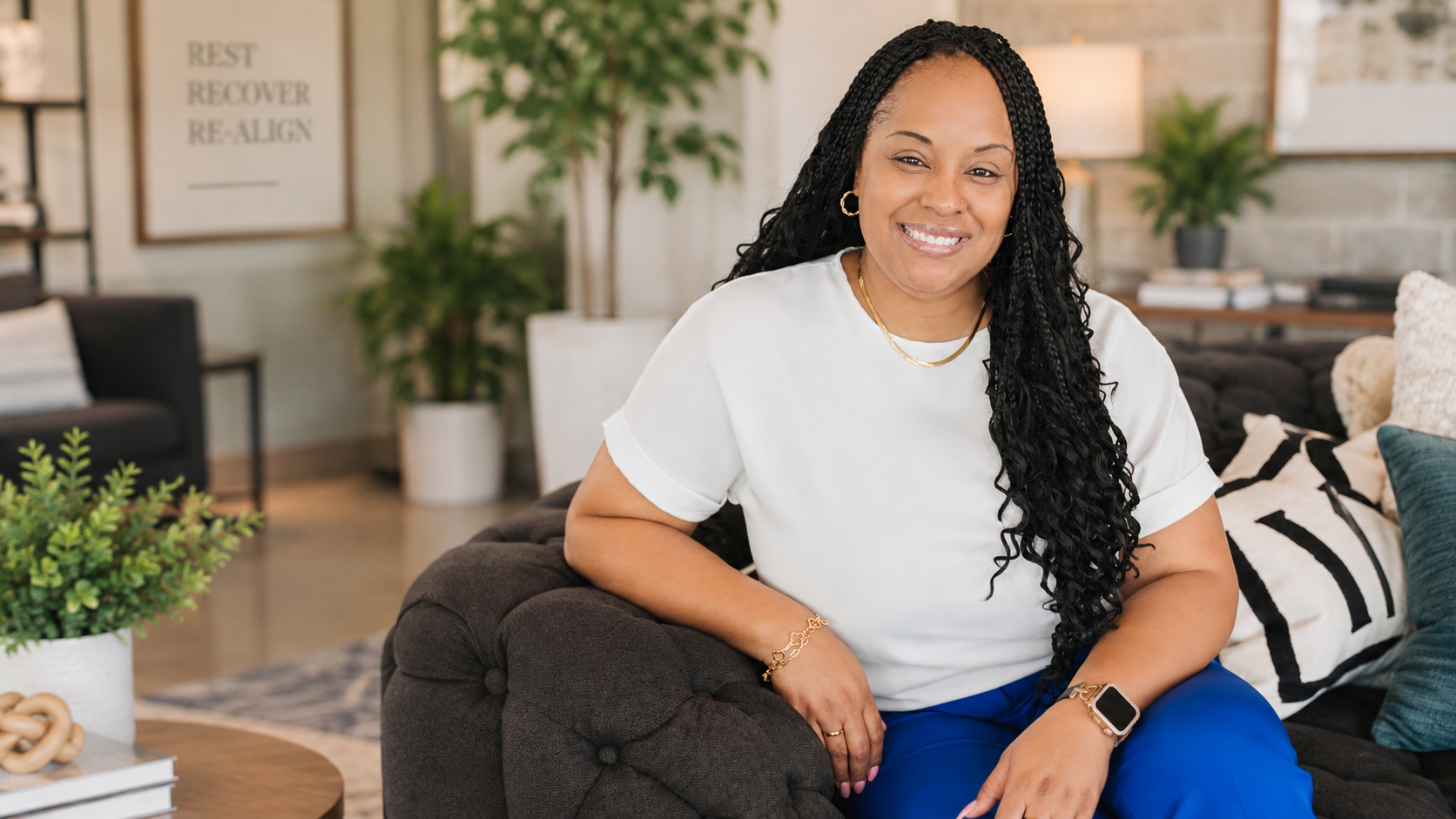 A woman with long curly black hair smiling while sitting on a dark gray sofa in a modern living room with plants, pillows, and artwork in the background.
