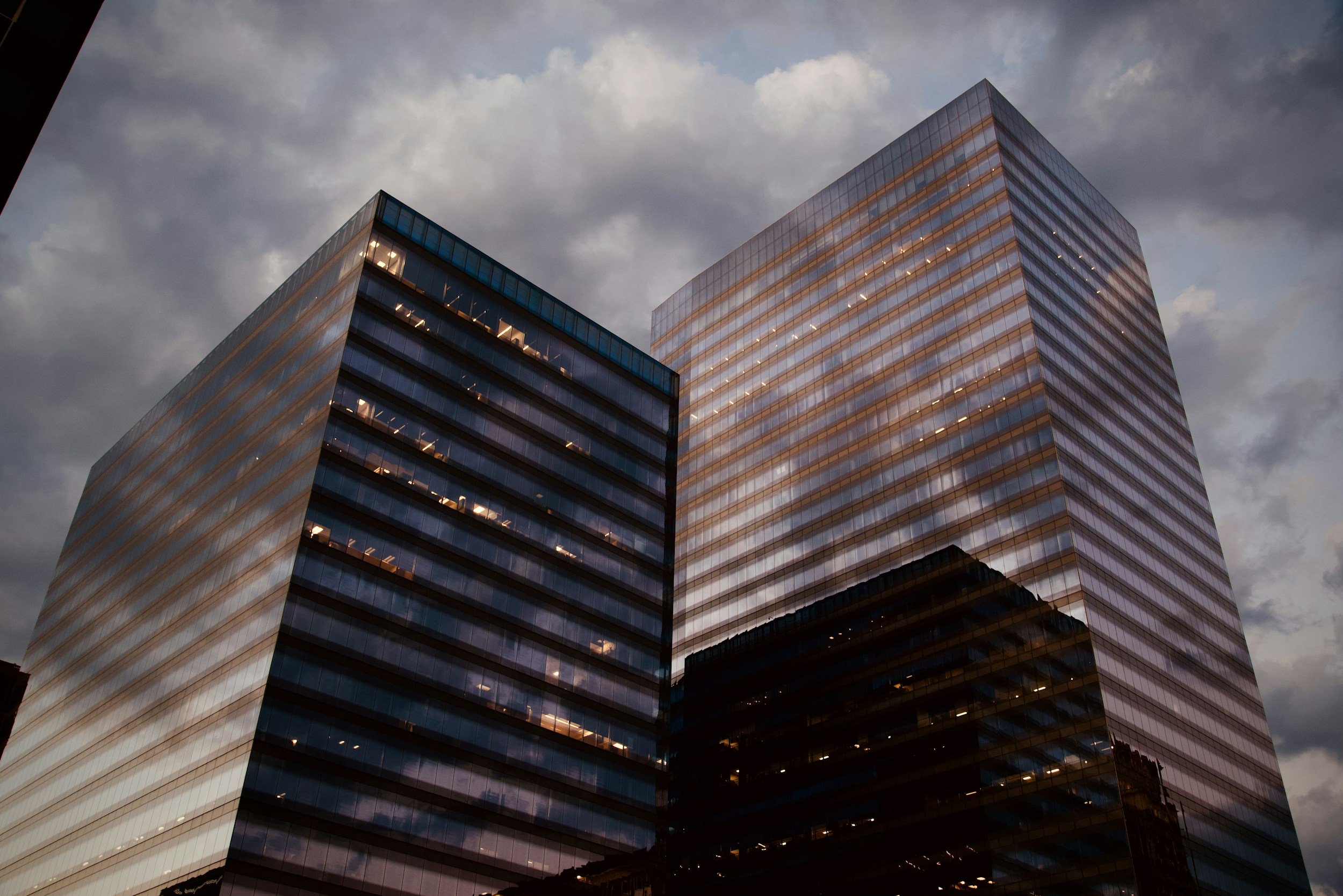 Zwei moderne Bürohochhäuser aus Glas vor bewölktem Himmel bei Dämmerung, reflektieren das Licht und die Wolken auf ihrer Fassade.