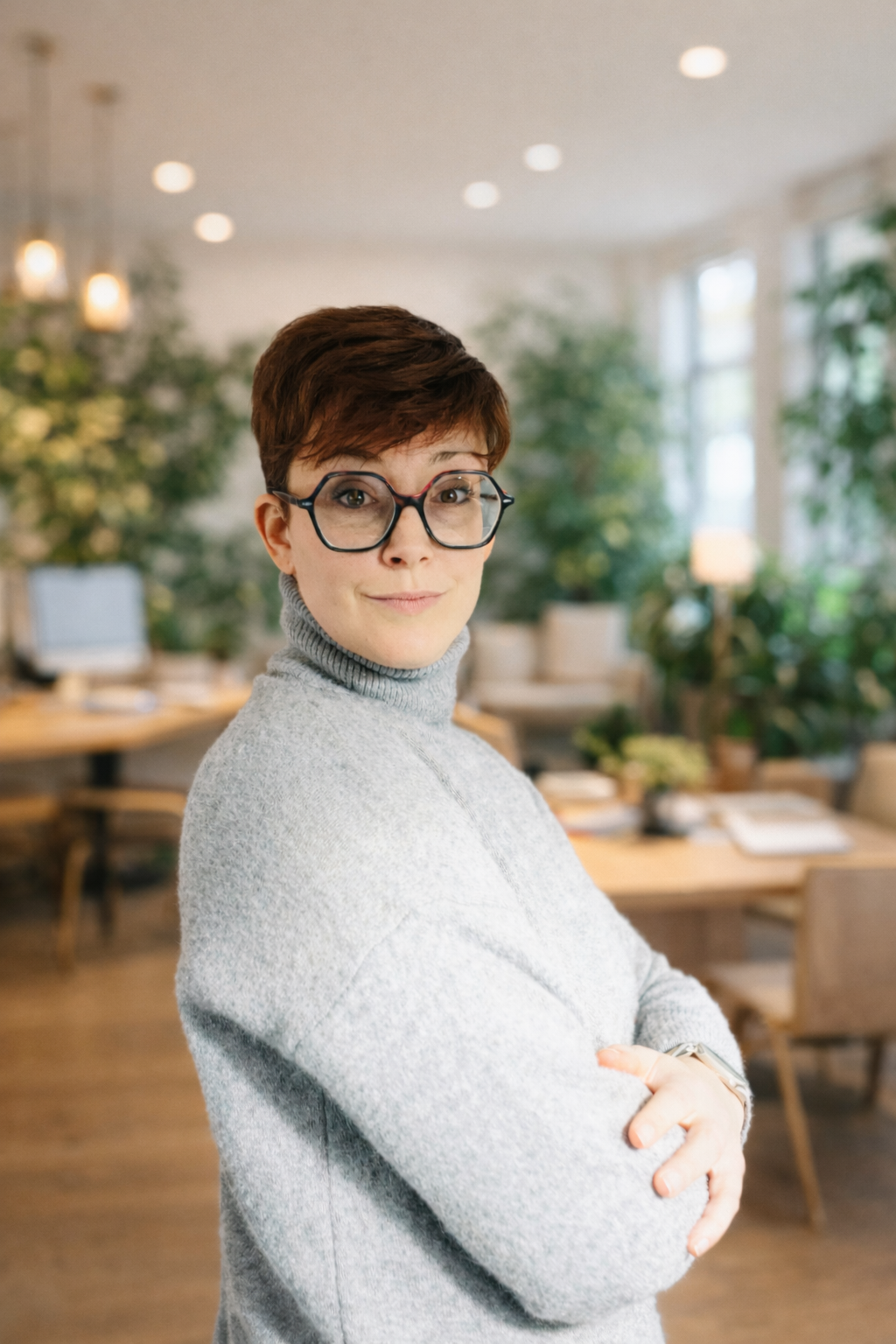Une femme avec des lunettes, portant un pull gris, dans un environnement intérieur lumineux avec des plantes et des meubles en bois.