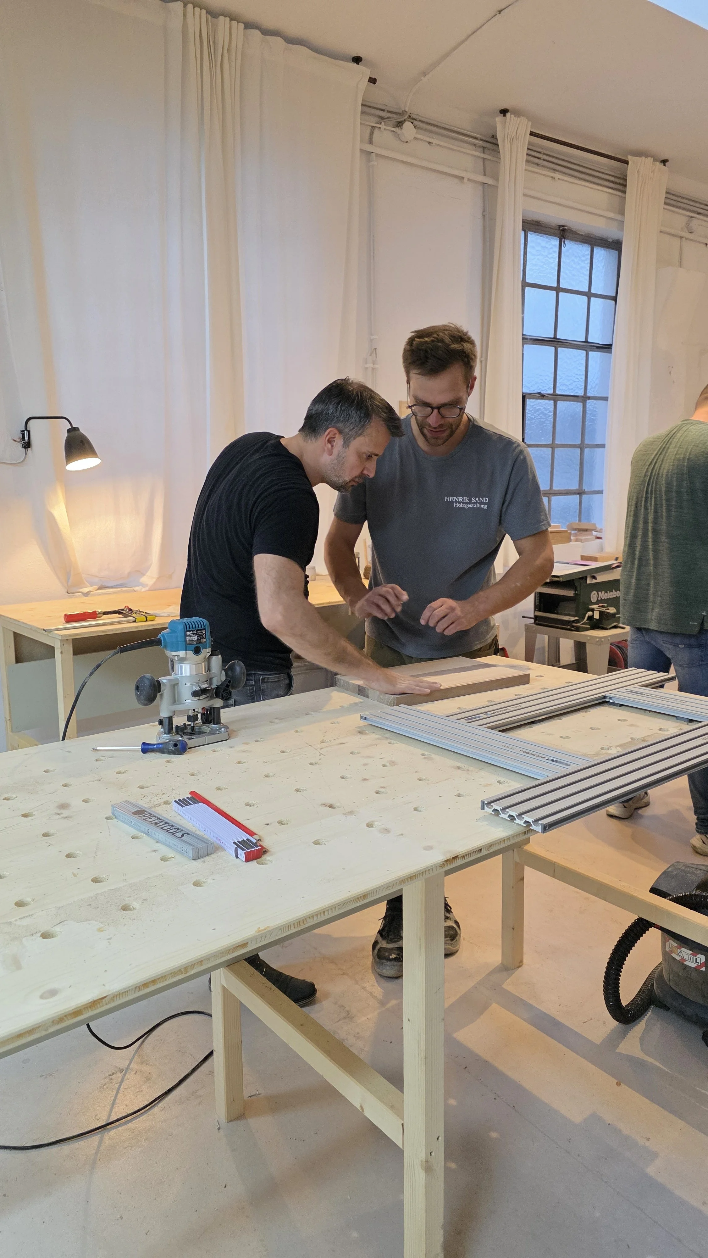 Zwei Männer arbeiten in einer Werkstatt an einem Holzprojekt. Der Tisch ist mit Werkzeugen und Aluminiumprofilen ausgestattet.