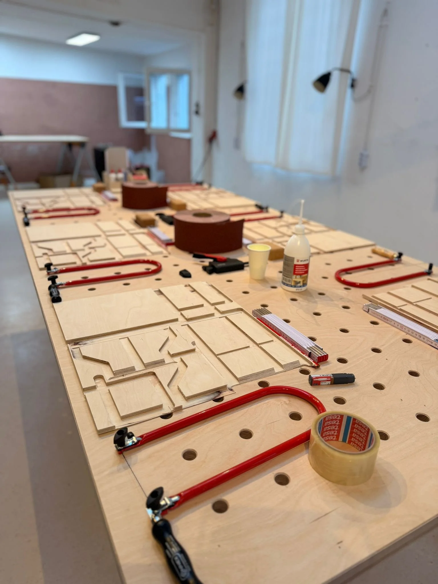 Ein Arbeitstisch mit Holzstücken, Werkzeugen und Messgeräten, vermutlich für einen Holzbau- oder Bastelauftrag in einem Werkraum.