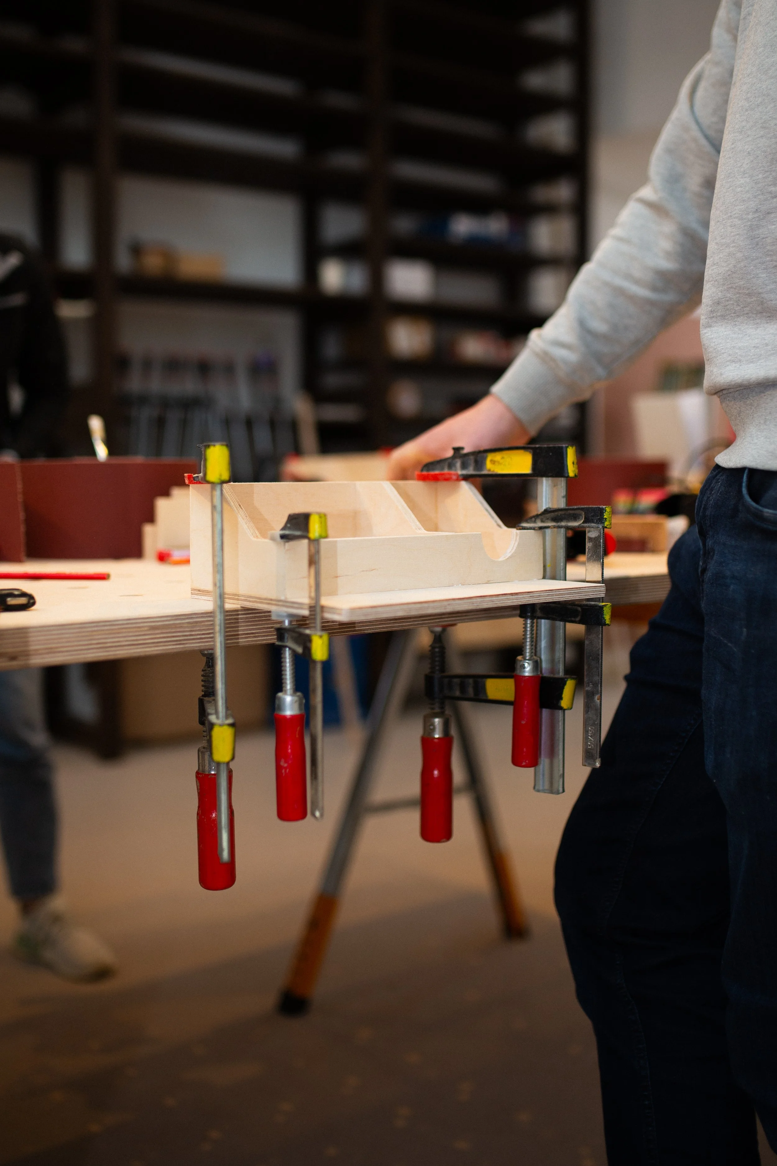Person, die an einem Holzstück in einer Werkstatt arbeitet, mit mehreren Schraubzwingen fixiert.