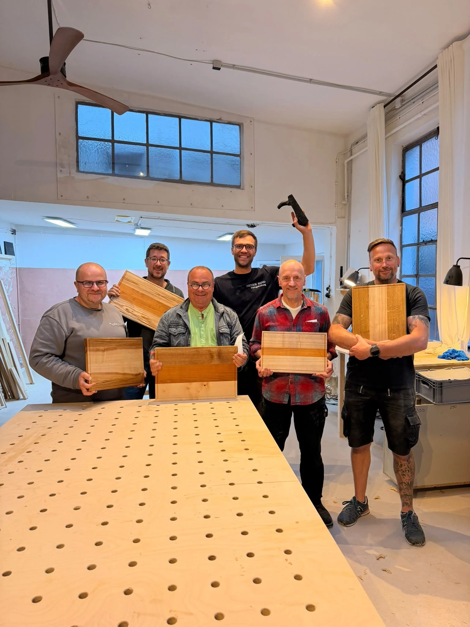 Eine Gruppe von sechs Männern steht in einem Raum, in dem Holzteile für Möbel hergestellt werden. Sie halten Holzbretter und Werkzeuge, lachen und posieren für das Foto.