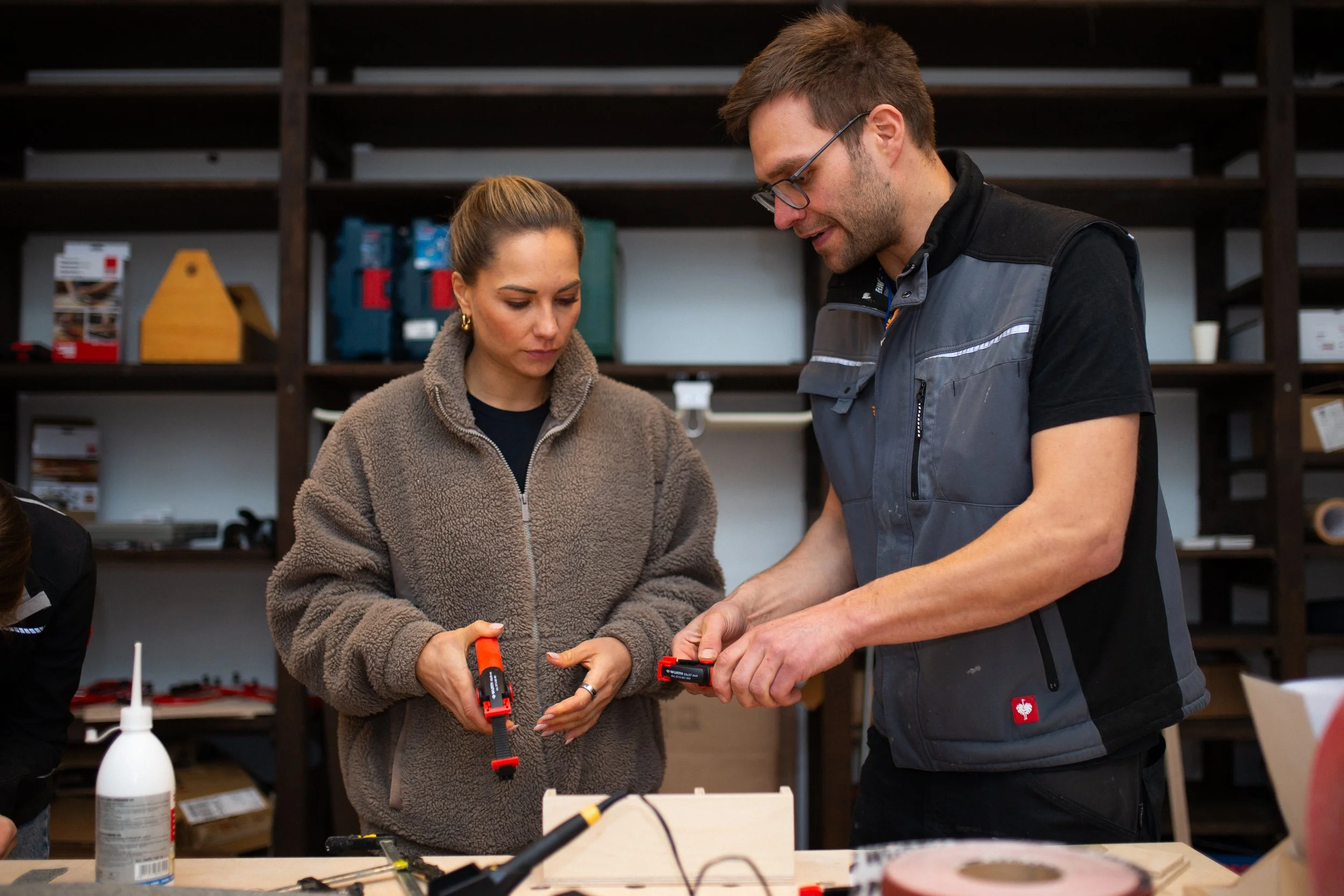 Zwei Menschen arbeiten in einer Werkstatt mit Werkzeugen, eine Frau schaut zu, der Mann erklärt etwas.