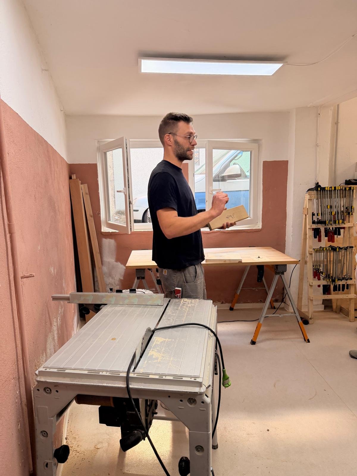 Ein Mann steht in einem Handswerksraum, hält ein Holzstück in der Hand und liest in einem Buch. Der Raum ist für Möbelbau eingerichtet, mit Werkzeugen und Werkbänken.
