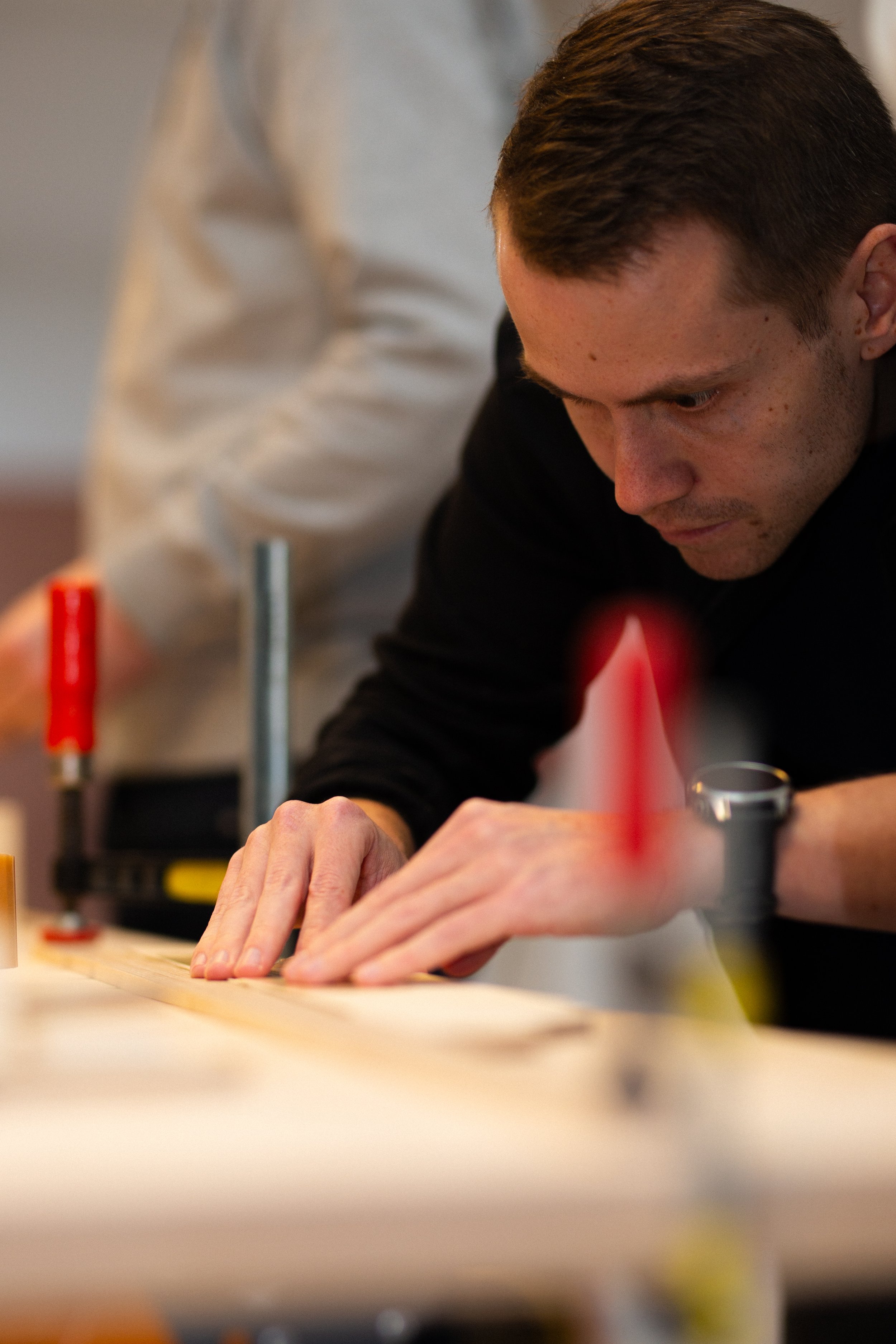 Ein junger Mann arbeitet konzentriert an einem Holzprojekt, während eine andere Person im Hintergrund hilft.