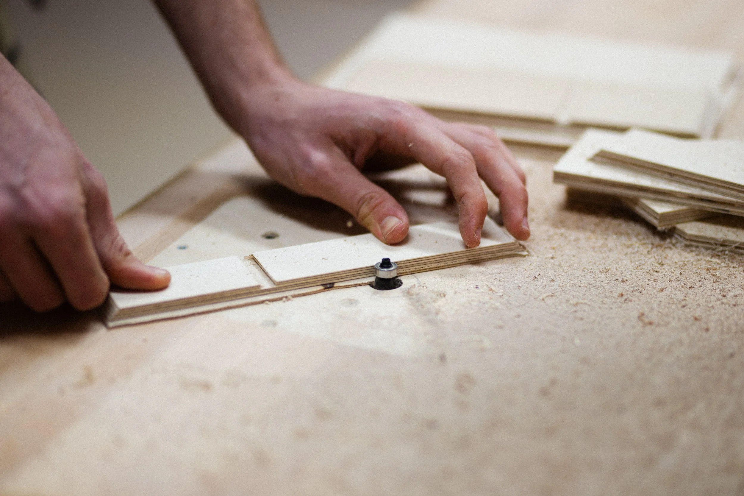 Hand legt Holzstück auf eine Werkbank, Werkzeuge und Holzstücke sind sichtbar.