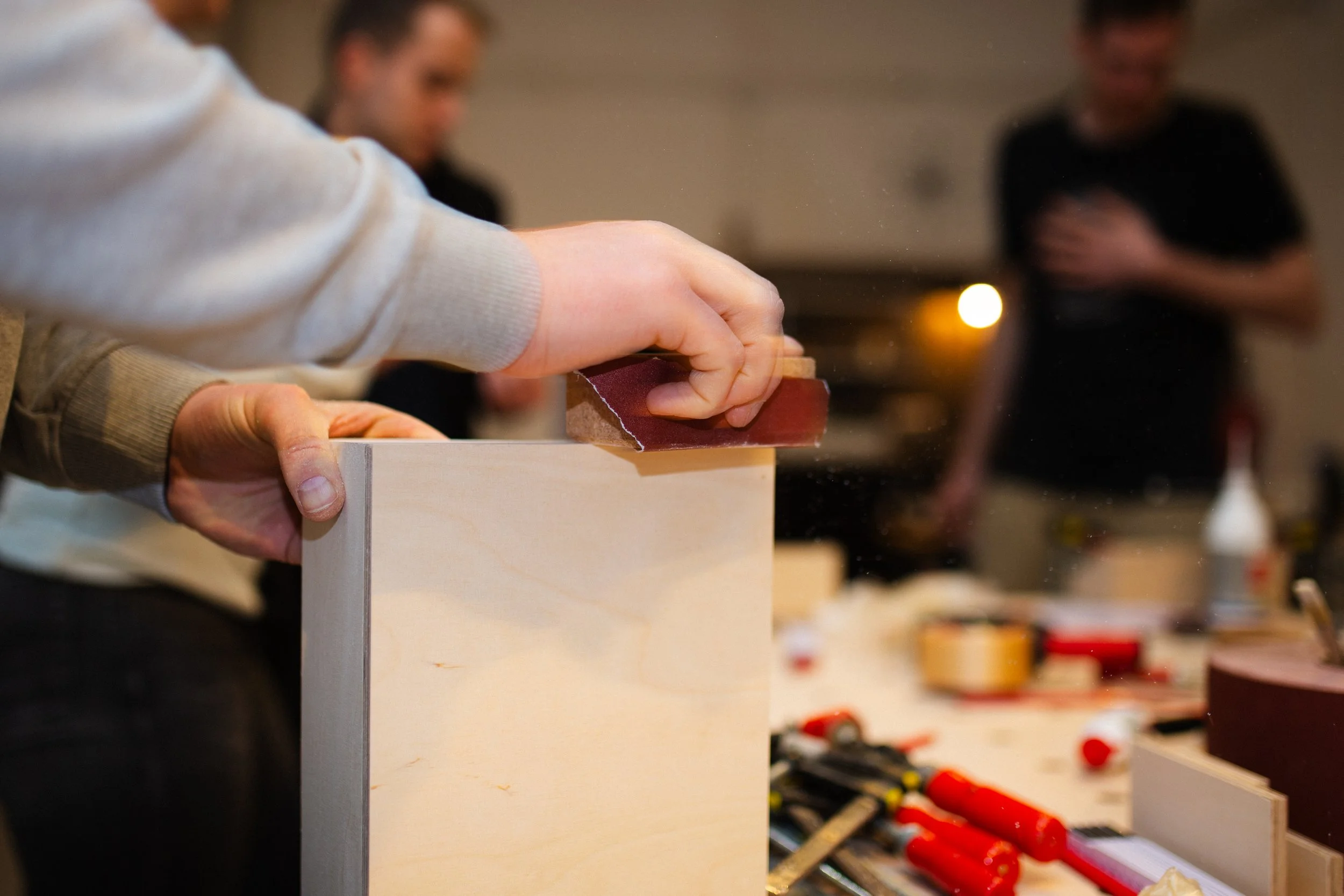 Person schleift die Kante eines Stück Holzes mit einem Schleifblock bei einem Workshop. Weitere Werkzeuge und Holzstücke liegen auf dem Tisch, im Hintergrund sind zwei weitere Menschen zu sehen.