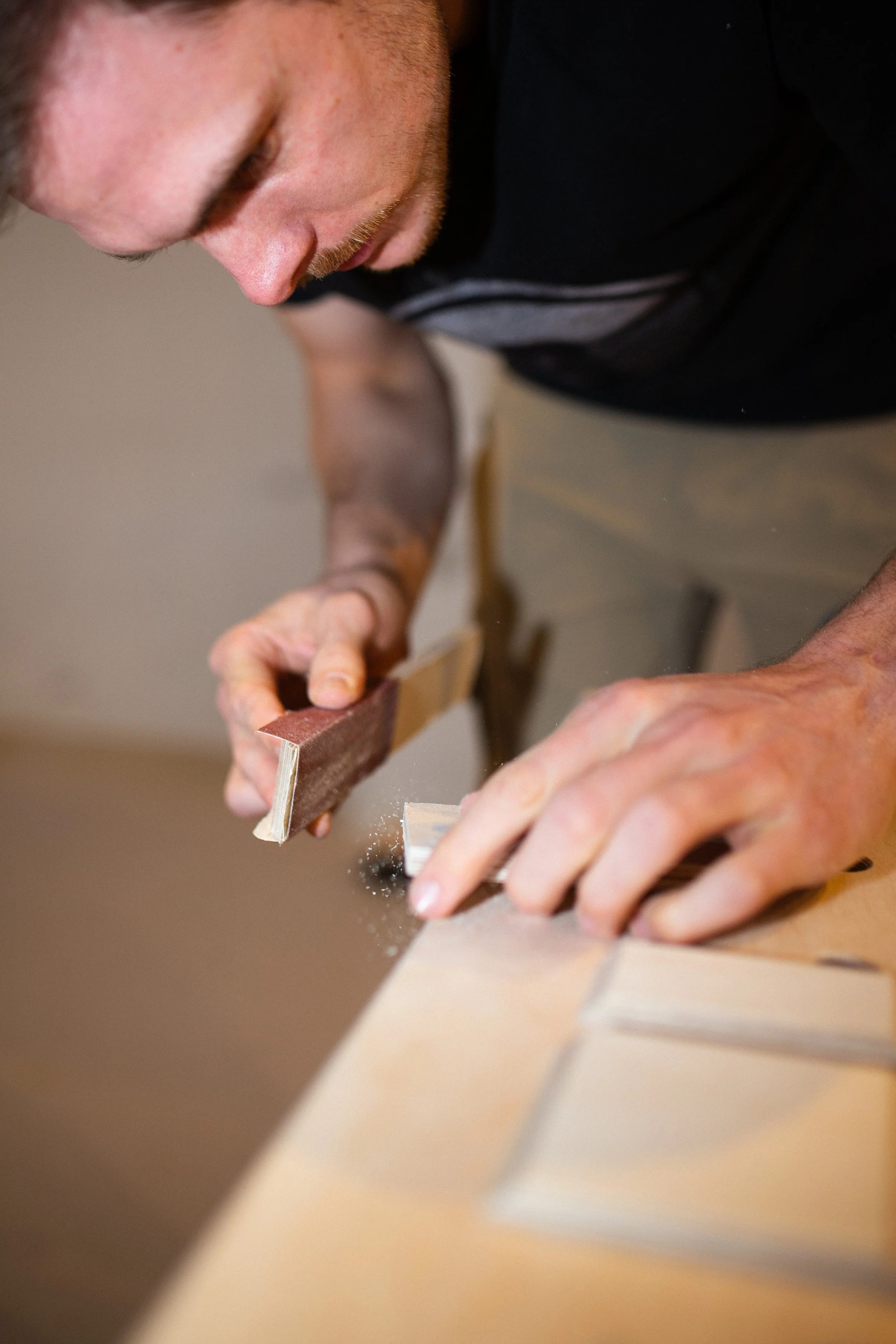 Ein Mann schleift ein Stück Holz mit einer Schleifmaschine auf einer Werkbank im Innenraum.