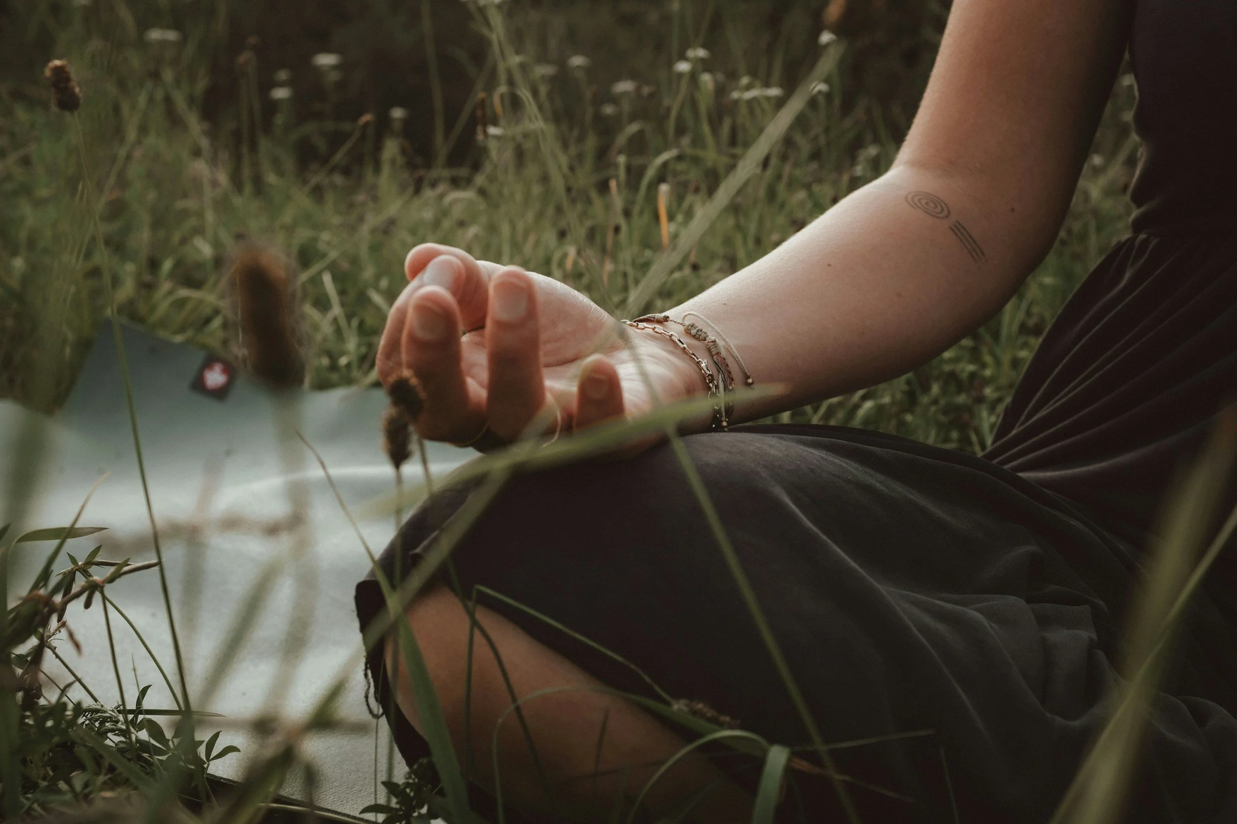 Person sitting cross-legged in a grassy field, with a tattoo on their arm and bracelets on their wrist, holding a card or paper, surrounded by tall grass.