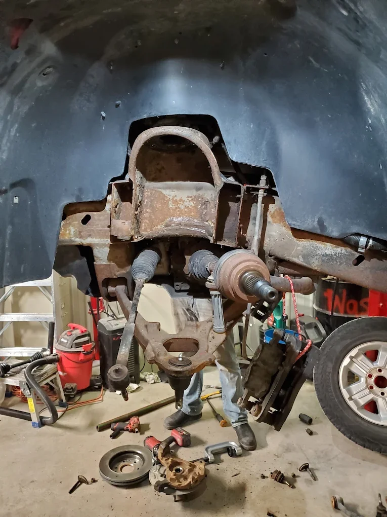 Rusty car suspension and brake assembly removed from a vehicle's wheel well, with tools and parts scattered on the garage floor.