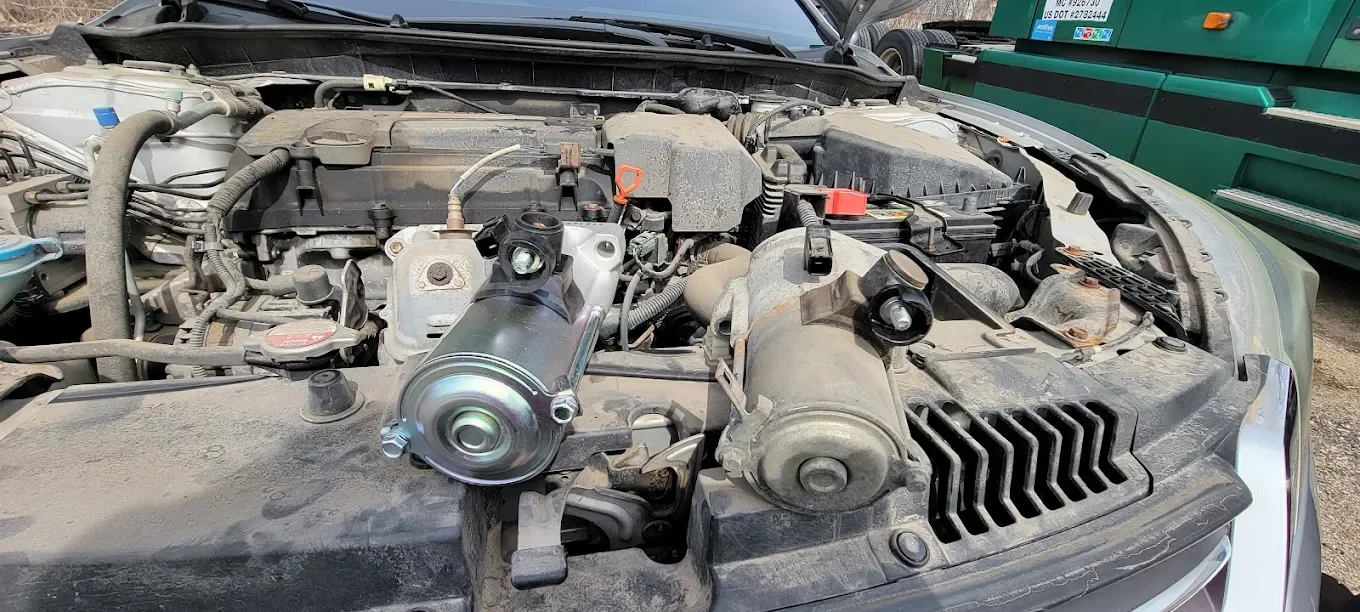 The engine bay of a car with visible various mechanical components, an aftermarket part mounted in the center, and dust and dirt accumulated on the engine surfaces.