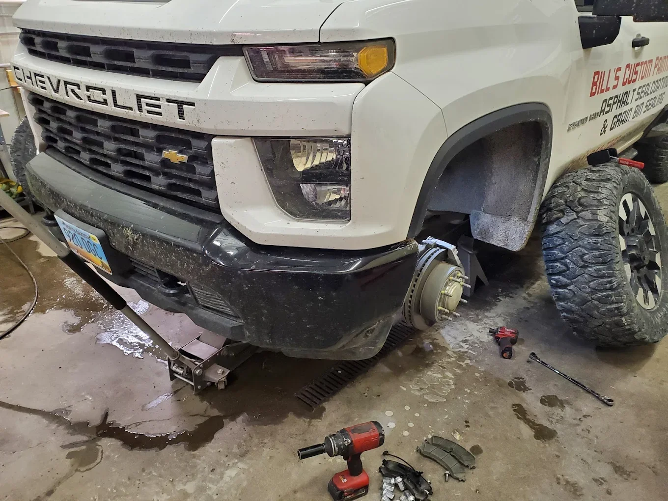 A white Chevrolet truck with front wheel removed, exposing brake system. Car parts, tools, and fluids are on the garage floor, indicating repair work.