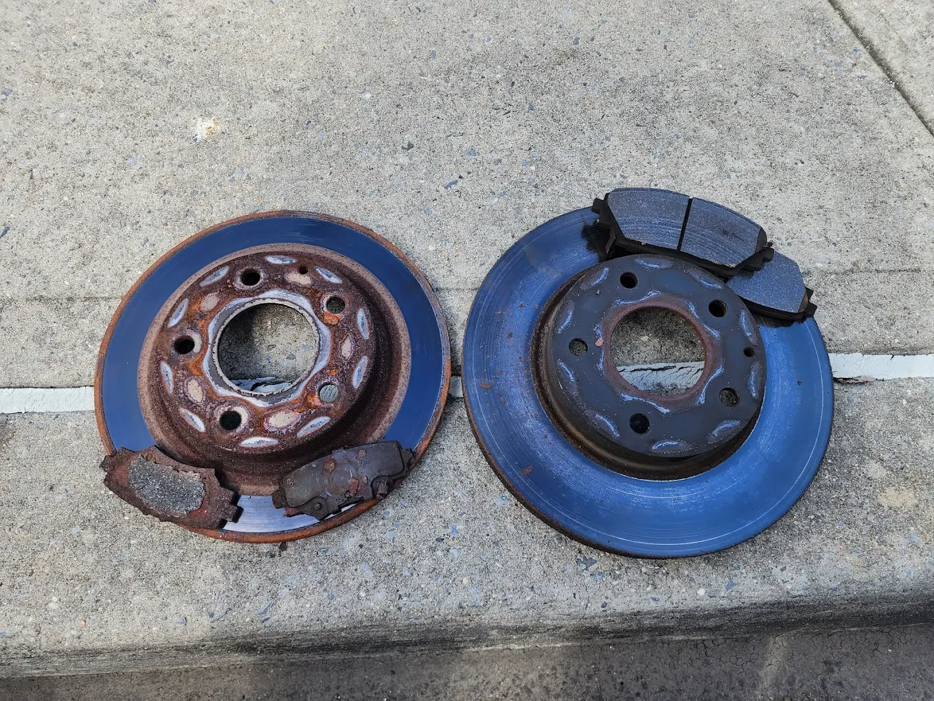 A pair of rusty and worn brake rotors with brake pads attached, lying on a concrete sidewalk.