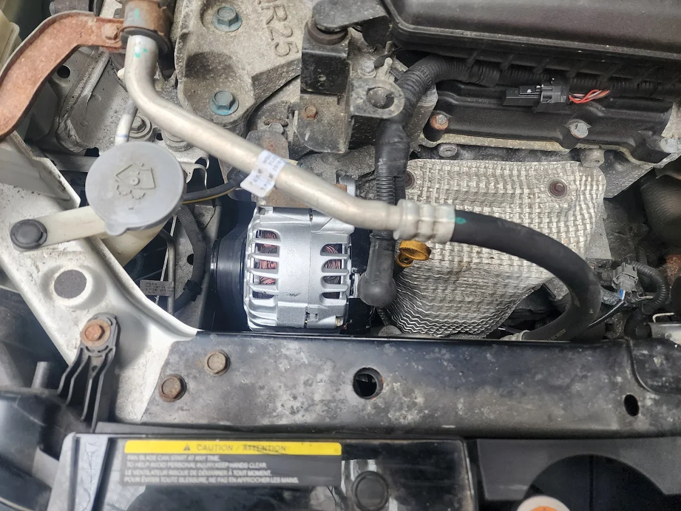 Close-up view of a car engine bay showing an alternator, various hoses, wiring, and metal components, with some rust and dirt visible.