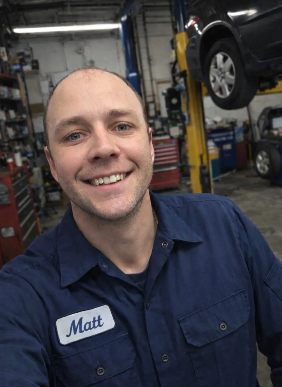 A smiling mechanic named Matt taking a selfie in an auto repair shop with cars and tools in the background.