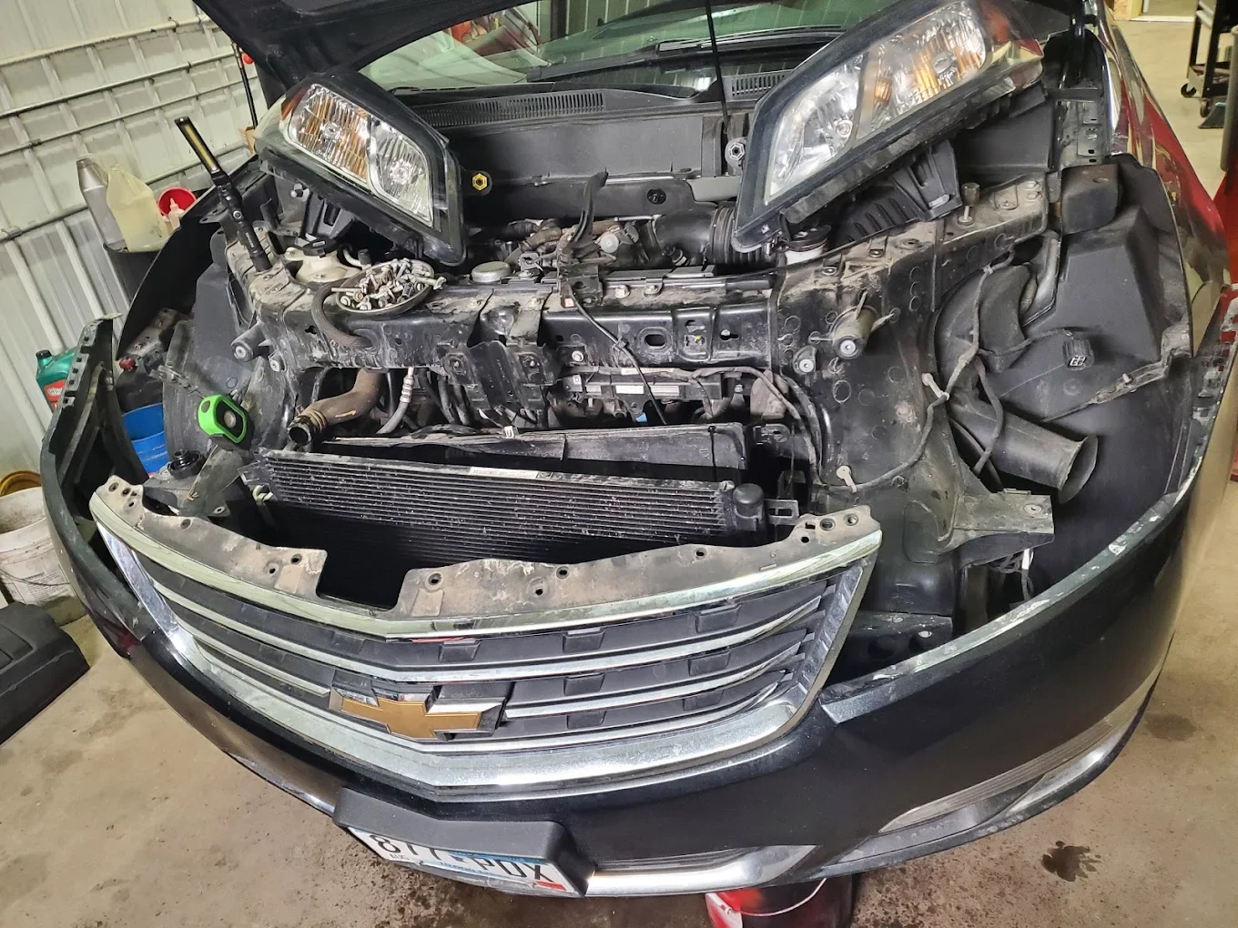 Front of a black Chevrolet car with the hood open, showing the engine compartment with parts removed and some tools around, inside a garage.