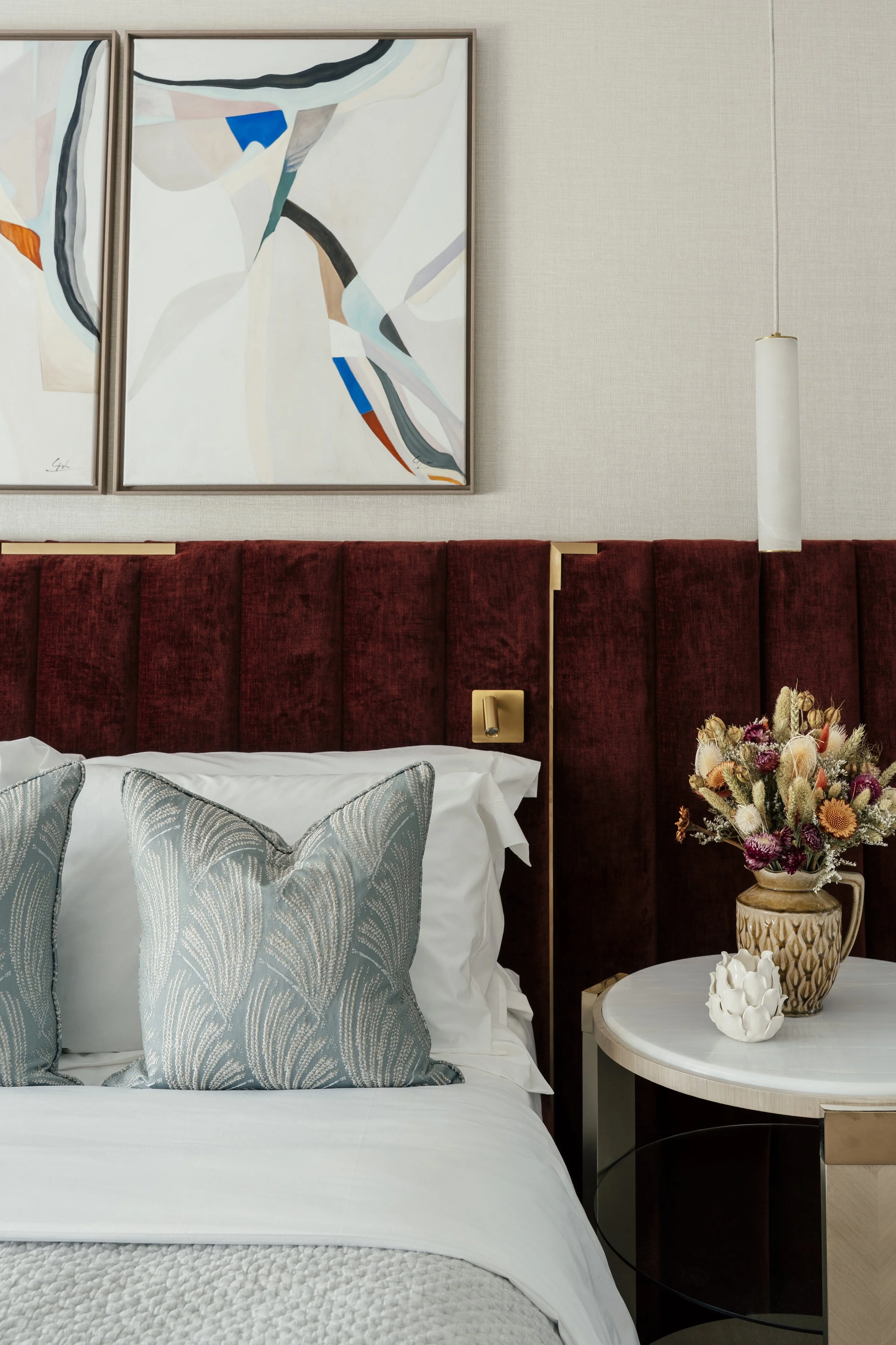 A bedroom with a white bed, two decorative pillows with leaf patterns, a red velvet headboard, a round side table with a vase of flowers, a decorative white ceramic object, a hanging white pendant light, abstract artwork on the wall, and a beige textured wall.