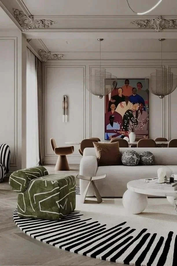 Modern living room with white walls, ornate crown molding, and large windows with curtains. Furniture includes a white sofa with brown pillows, a green armchair, a small white table, and a darker brown side table. Decor features a colorful abstract painting on the wall, a white vase with flowers, a striped black-and-white rug, and modern light fixtures hanging from the ceiling.