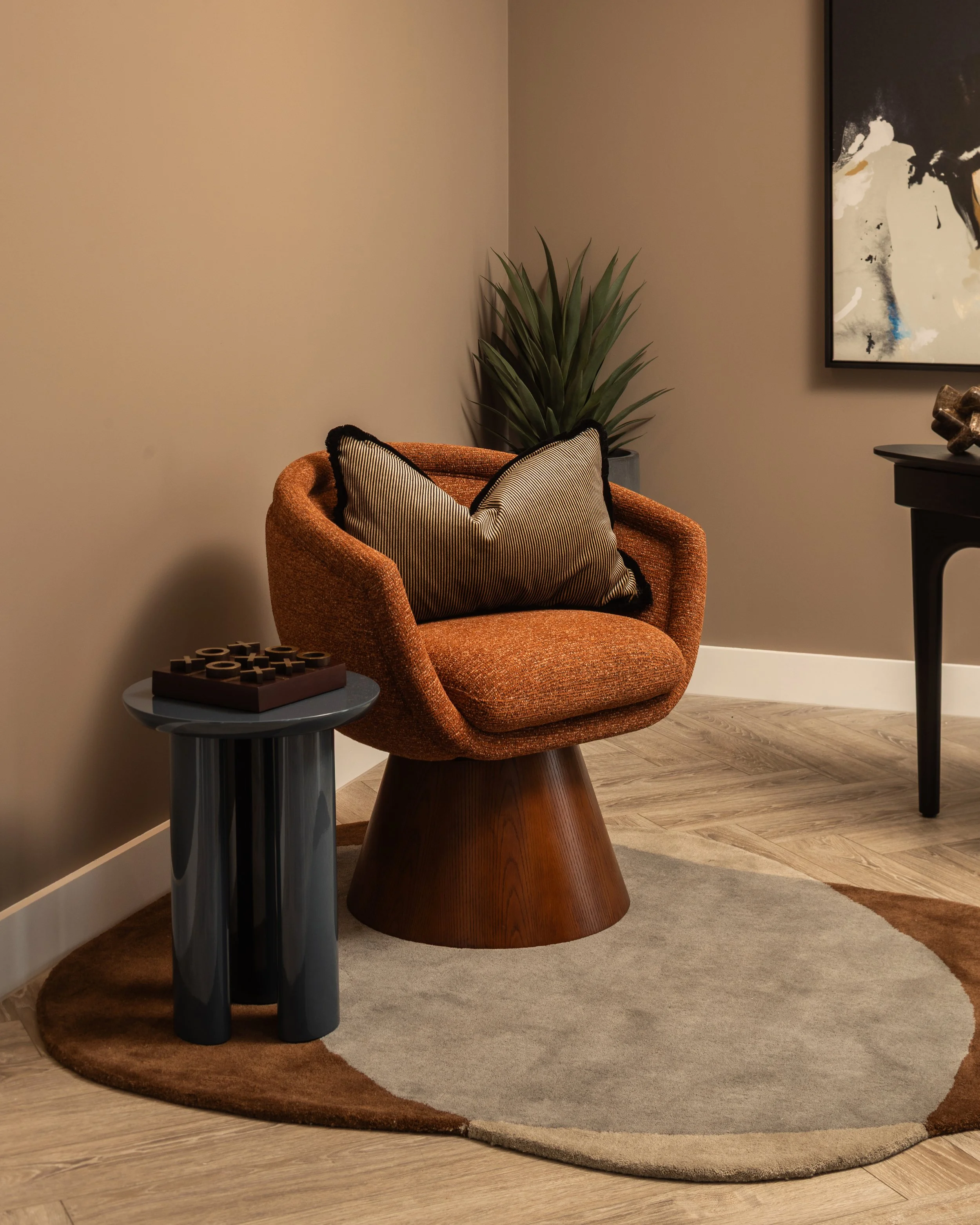An orange upholstered armchair with a striped pillow, standing on a round beige and brown rug, next to a small black side table with decorative items, with a tall green plant behind it and a black and white abstract painting on the wall.
