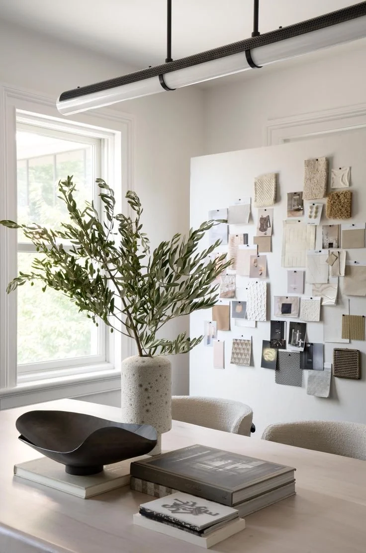 A modern interior with a white table holding a large ceramic vase with greenery, a black decorative bowl, and stacked books. In the background, a white wall features a gallery of fabric samples or swatches. There's a window bringing in natural light and a sleek ceiling light fixture.