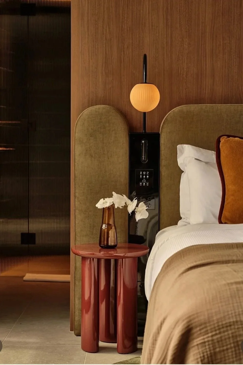 A bedroom with a bed, white open pillows, mustard-colored pillows, a red side table with white flowers in a brown vase, a wall-mounted bedside lamp, and a wood-paneled wall.