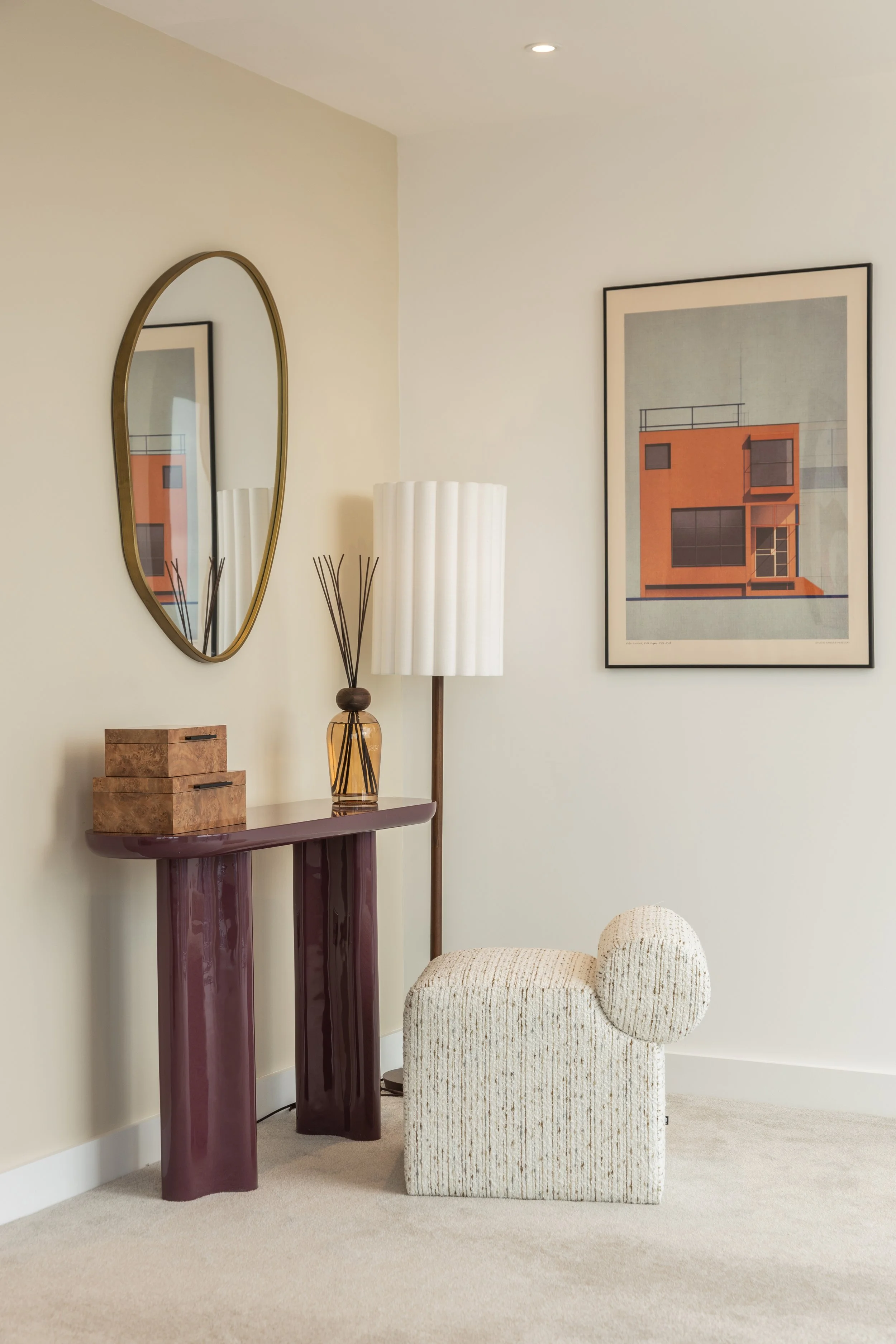 Cozy living room corner with a modern sculpture, a tall floor lamp with a white lampshade, a decorative mirror, a framed artwork of a building, and a textured beige armchair.