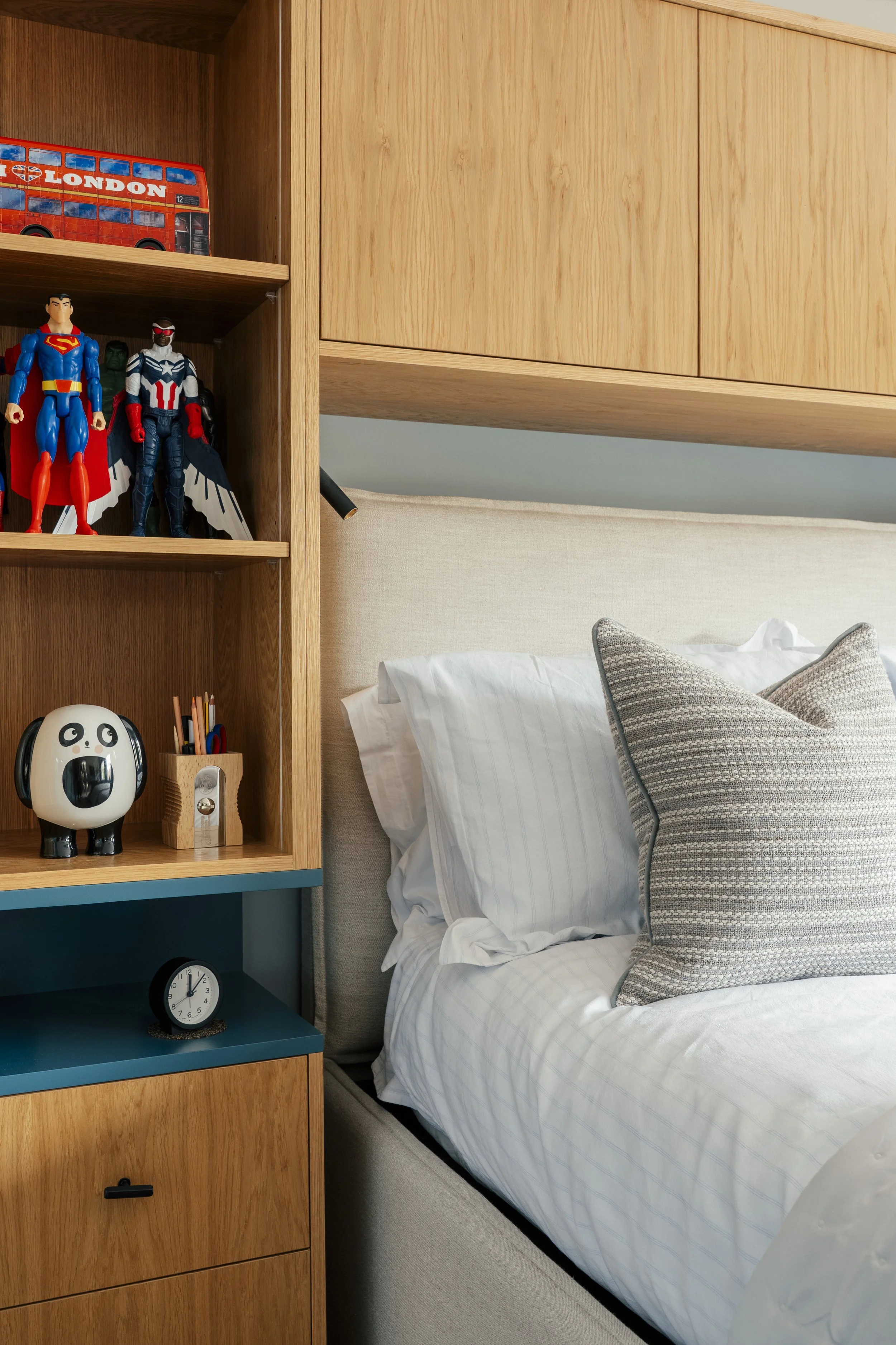 Close-up of a bed with pillows and a headboard, near a wooden shelf with toys, a clock, and a lamp in a bedroom.