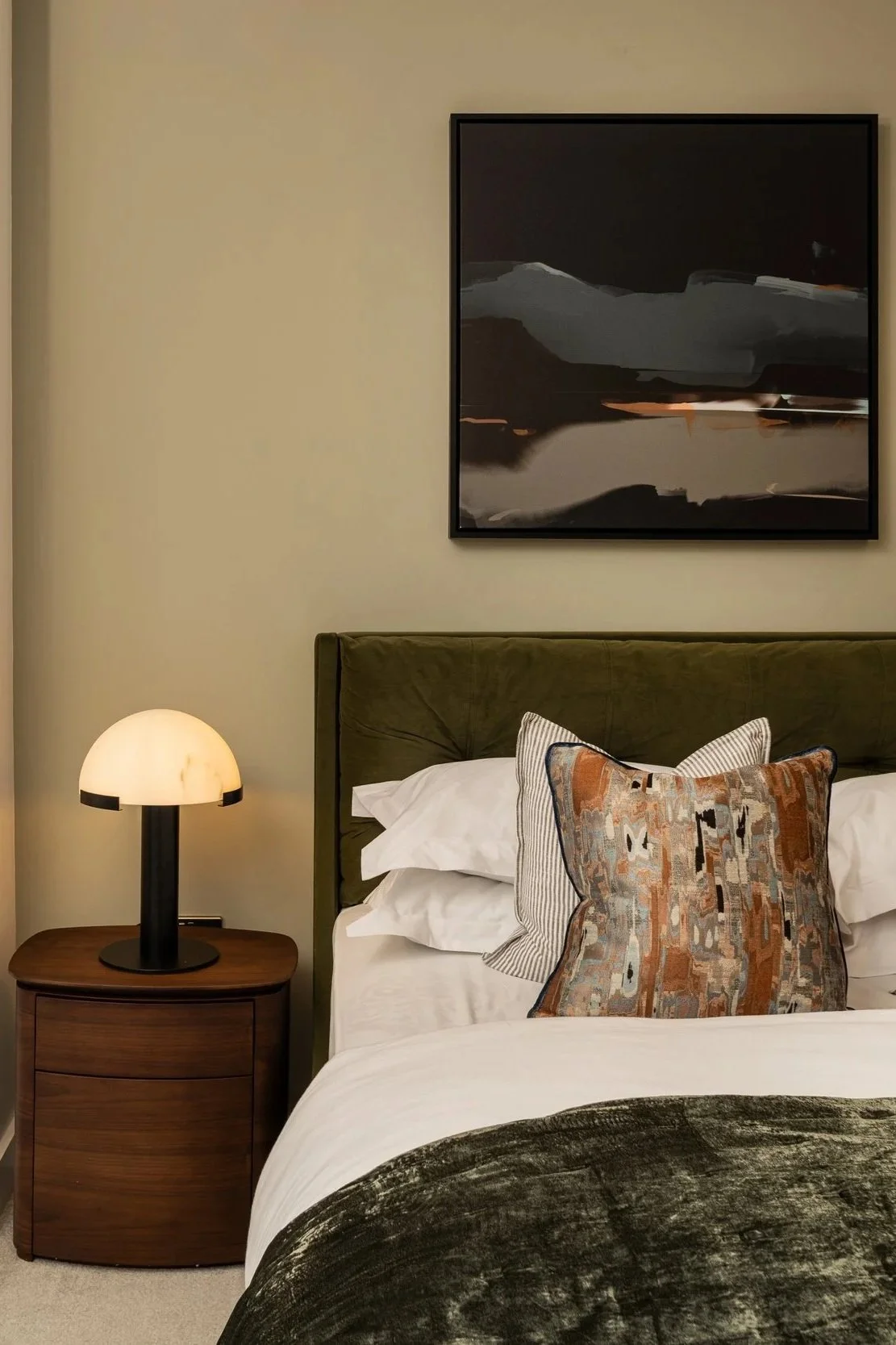 A bedroom with a bed having white sheets, pillows, and a decorative pillow. A wooden nightstand with a lamp and a dark abstract painting on the wall