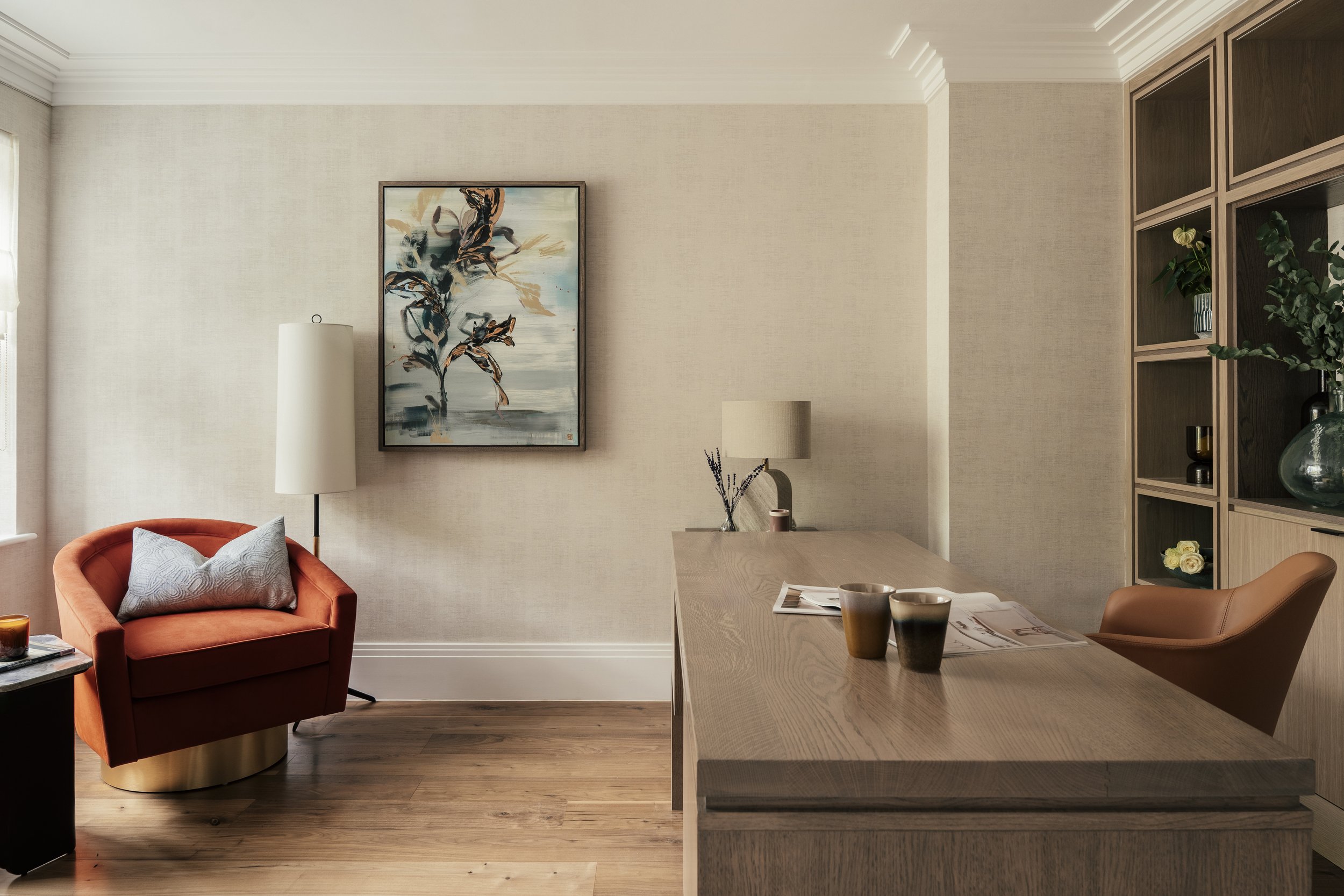 Contemporary living room with orange armchair, wooden desk, and shelving with plants and decorative items, beige walls, and hardwood flooring.