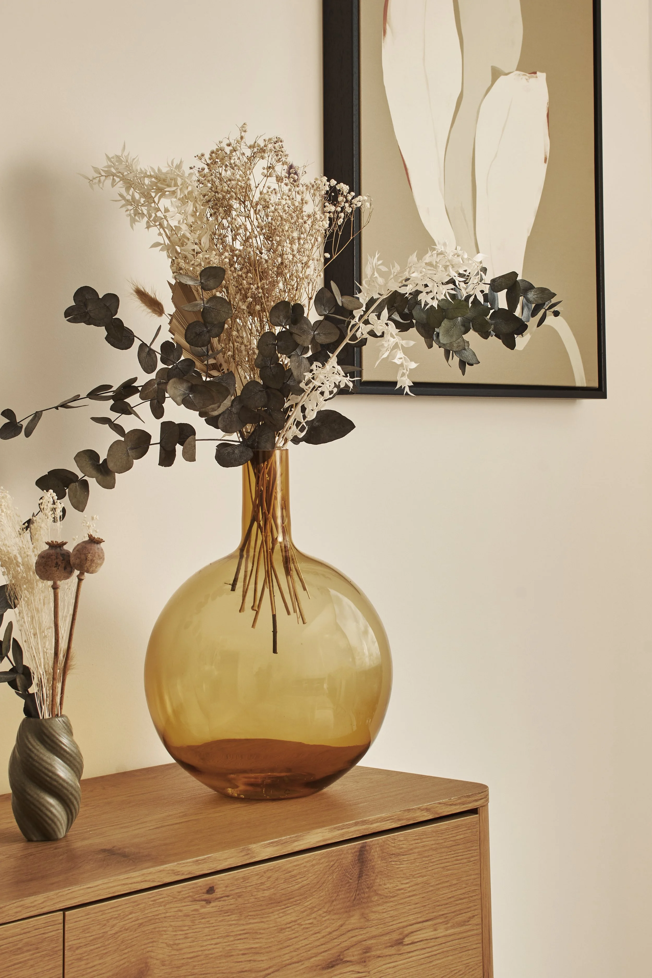 A large amber glass vase with dried flowers on a wooden surface, with a smaller gray vase with dried flowers nearby and a framed abstract art piece on a wall behind.
