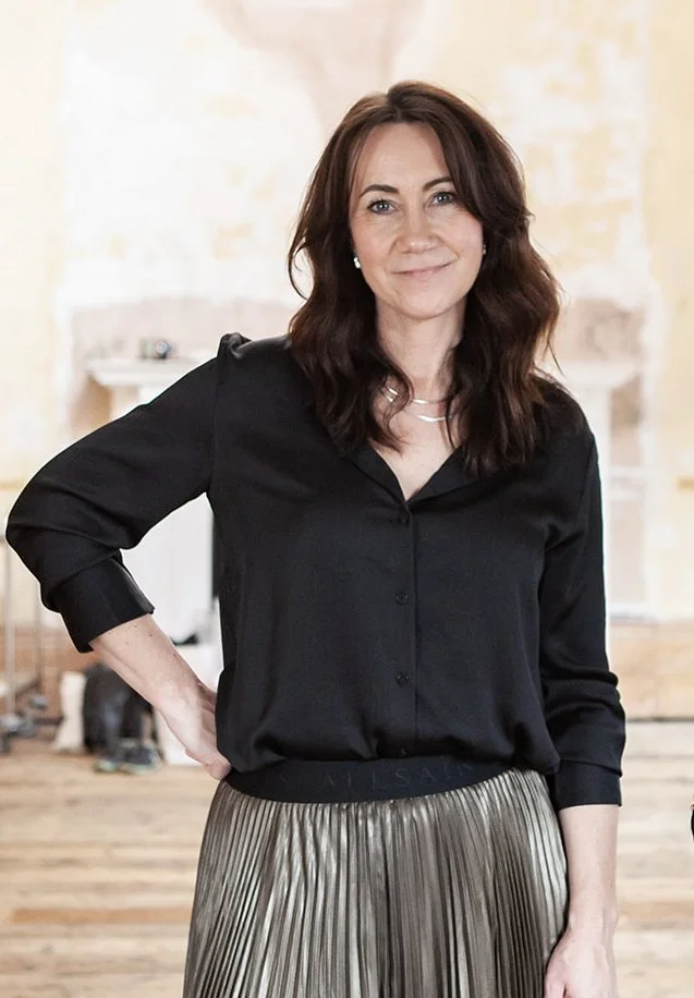 Woman with shoulder-length brown hair in a black blouse and metallic pleated skirt standing indoors with her hand on her hip and smiling.