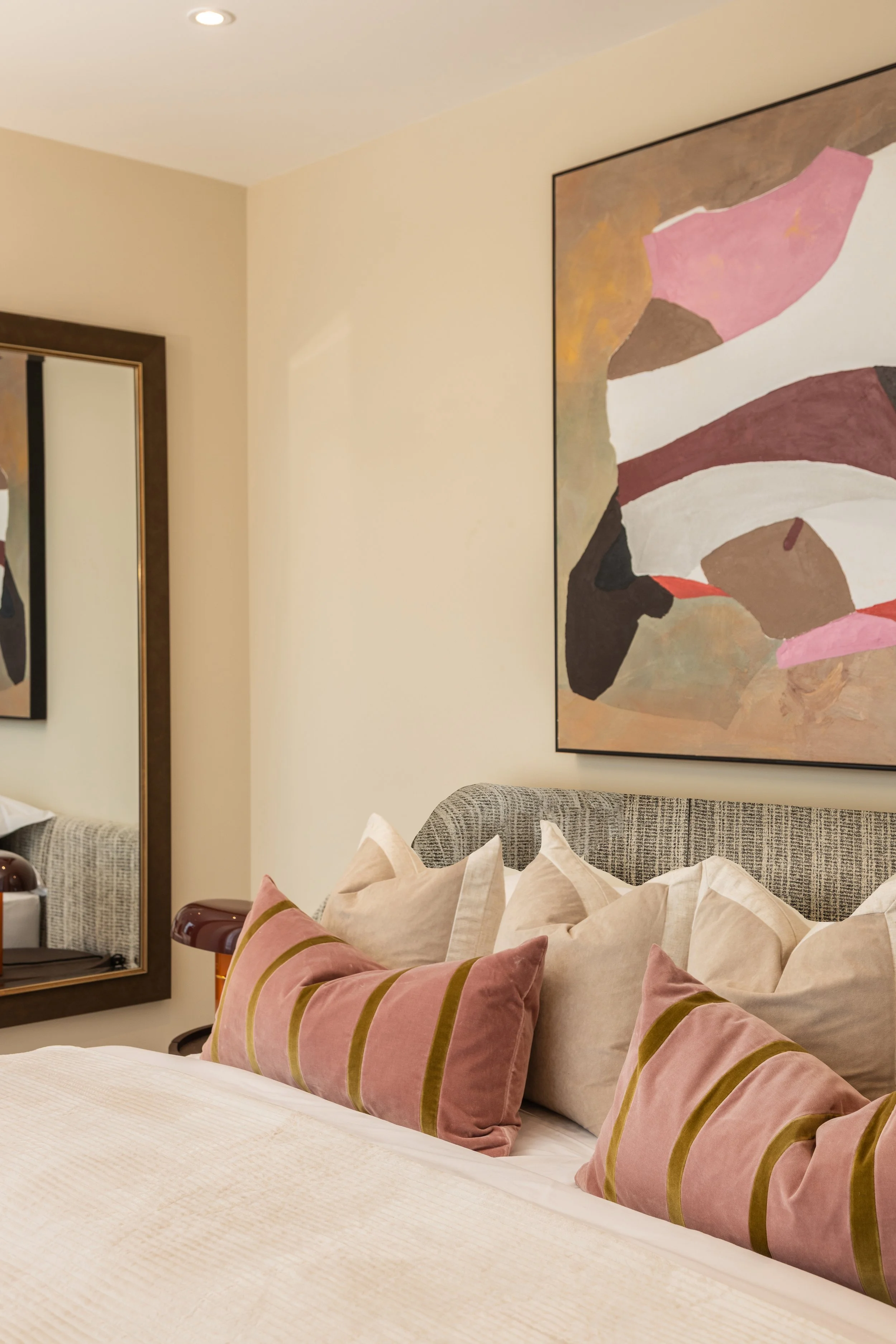 A bedroom with a bed featuring multiple pillows, a framed standing mirror, and abstract artwork on the wall.