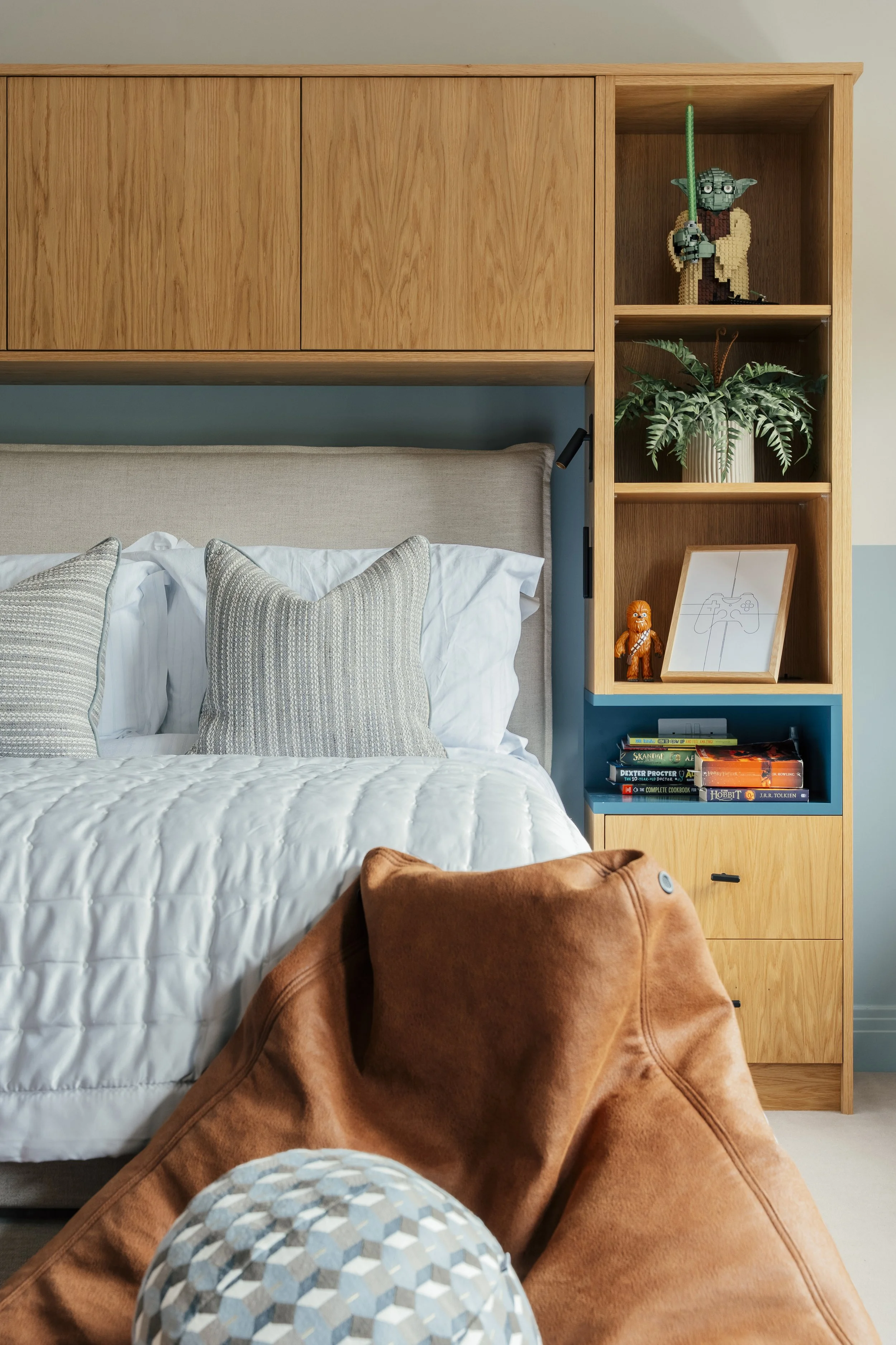 A cozy bedroom with a bed featuring white bedding and two plaid pillows. Beside the bed is a built-in wooden shelving unit with decorative items, books, and a framed drawing of a video game controller. In the foreground, there is a brown leather bean bag and a geometric-patterned ottoman.