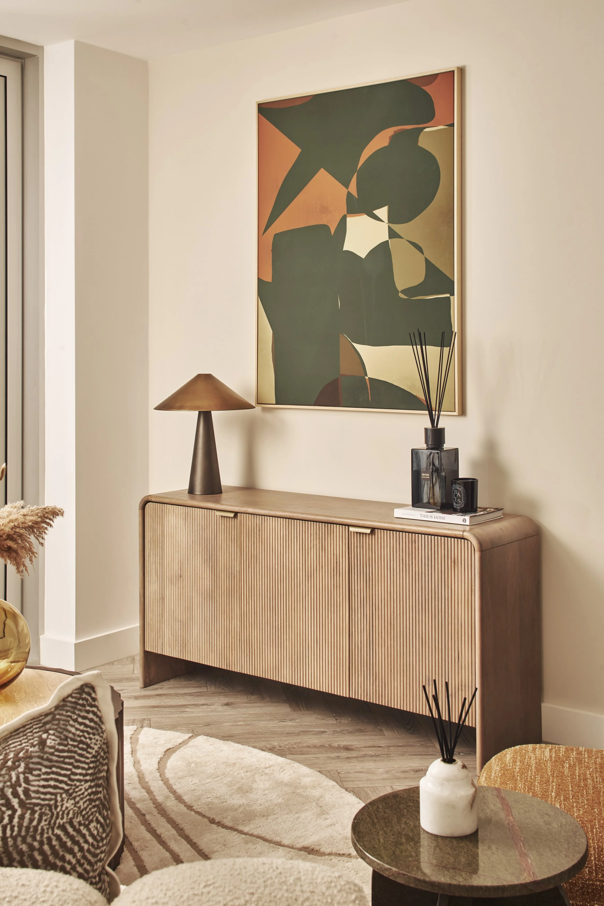 Modern living room interior with a wooden sideboard, abstract wall art, a table lamp, and decorative items like candles and reed diffusers.