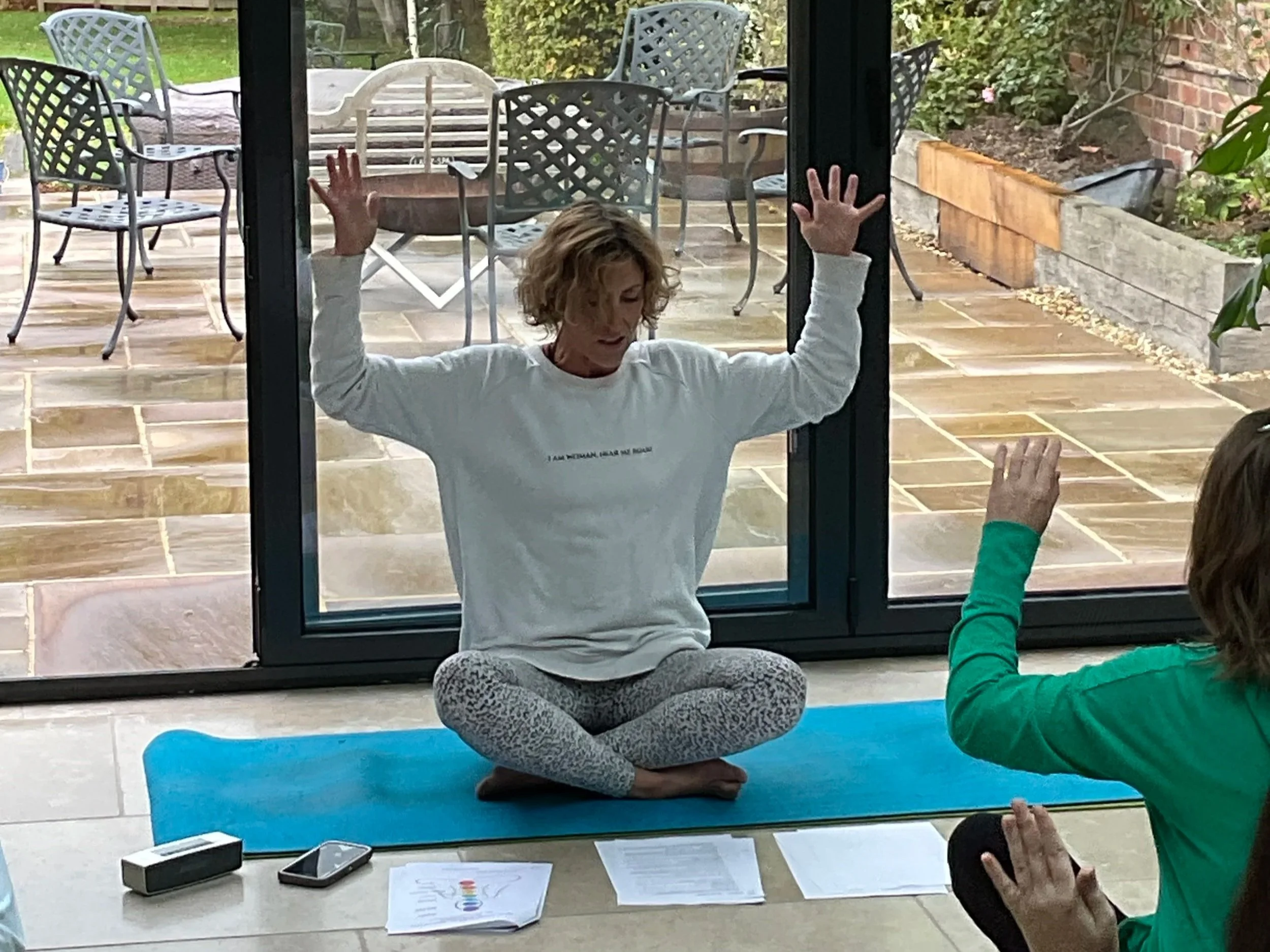 Woman practicing yoga in front of a glass door, seated cross-legged on a blue yoga mat, with her hands raised and palms open. Two smartphones and some papers are on the floor nearby. Another person, partially visible, is raising their hand.