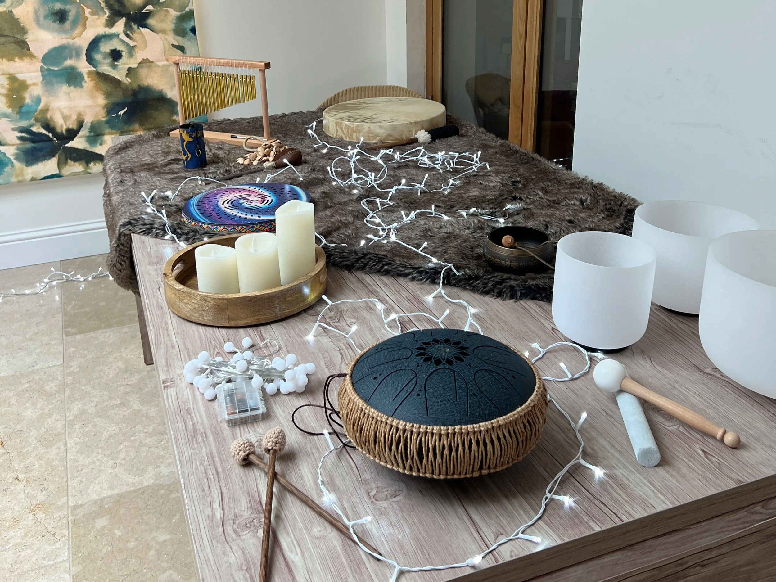 A wooden table with white LED string lights, ritual objects, candles, and singing bowls, set on a cozy furry blanket with a colorful wall hanging in the background.