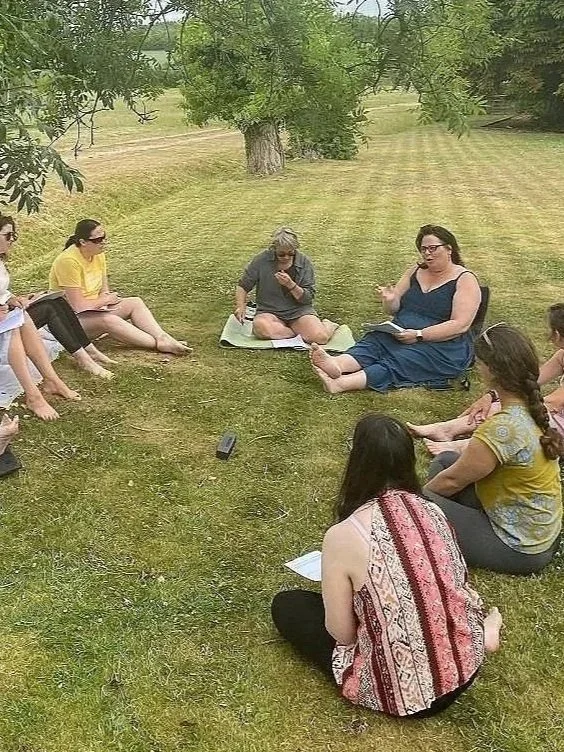 Group of women and girls sitting on grass in a circle outdoors, some taking notes and listening.