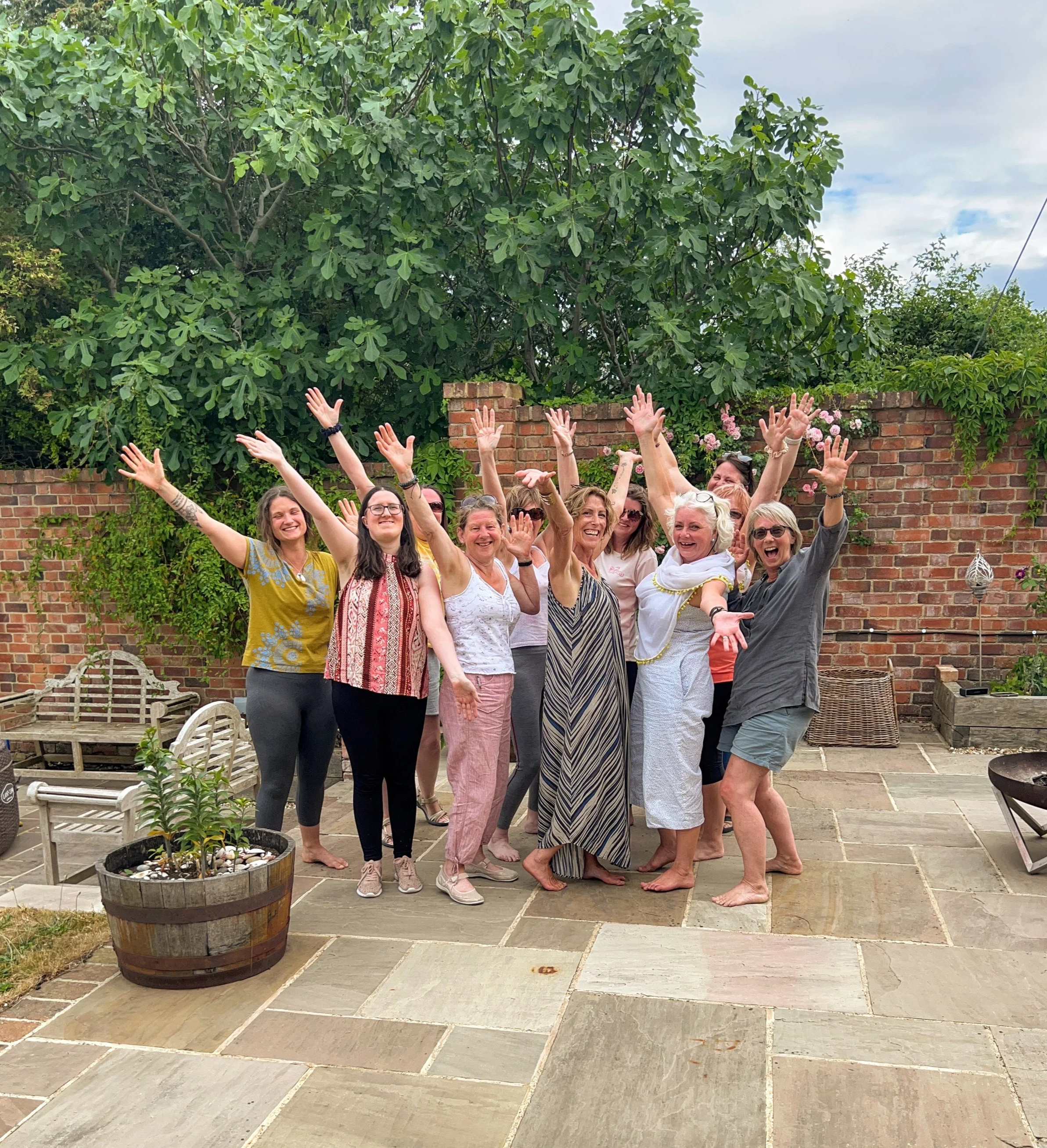 A group of women celebrating outdoors on a patio with their hands raised and smiling, with a large tree and a brick wall in the background.
